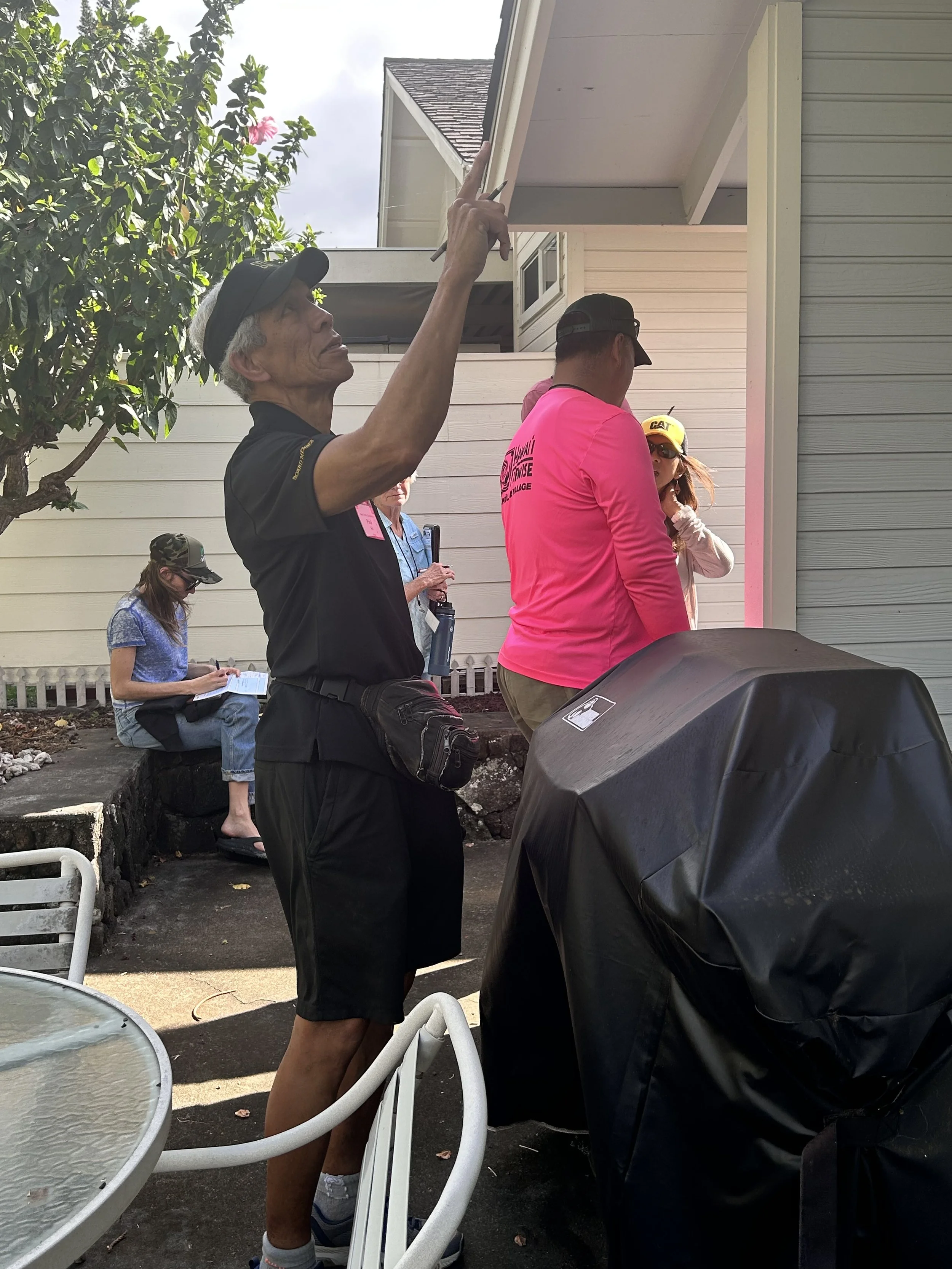A man in a black polo shirt and shorts is holding a pencil and pointing to the roof of a house during what appears to be a home inspection or evaluation. Several people are gathered around, with one woman sitting on a ledge and another woman on the p