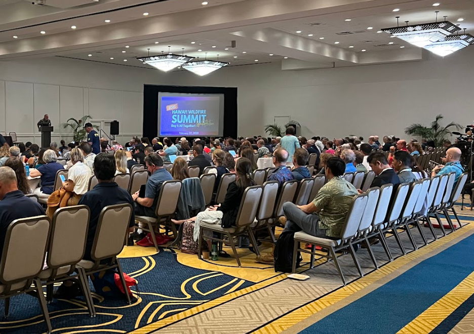 Large conference room filled with attendees seated facing a stage with a large screen. The screen displays the text 'Hawaii Wildfire Summit.' A speaker is at a podium on the stage, and the room is decorated with chandeliers and plants.