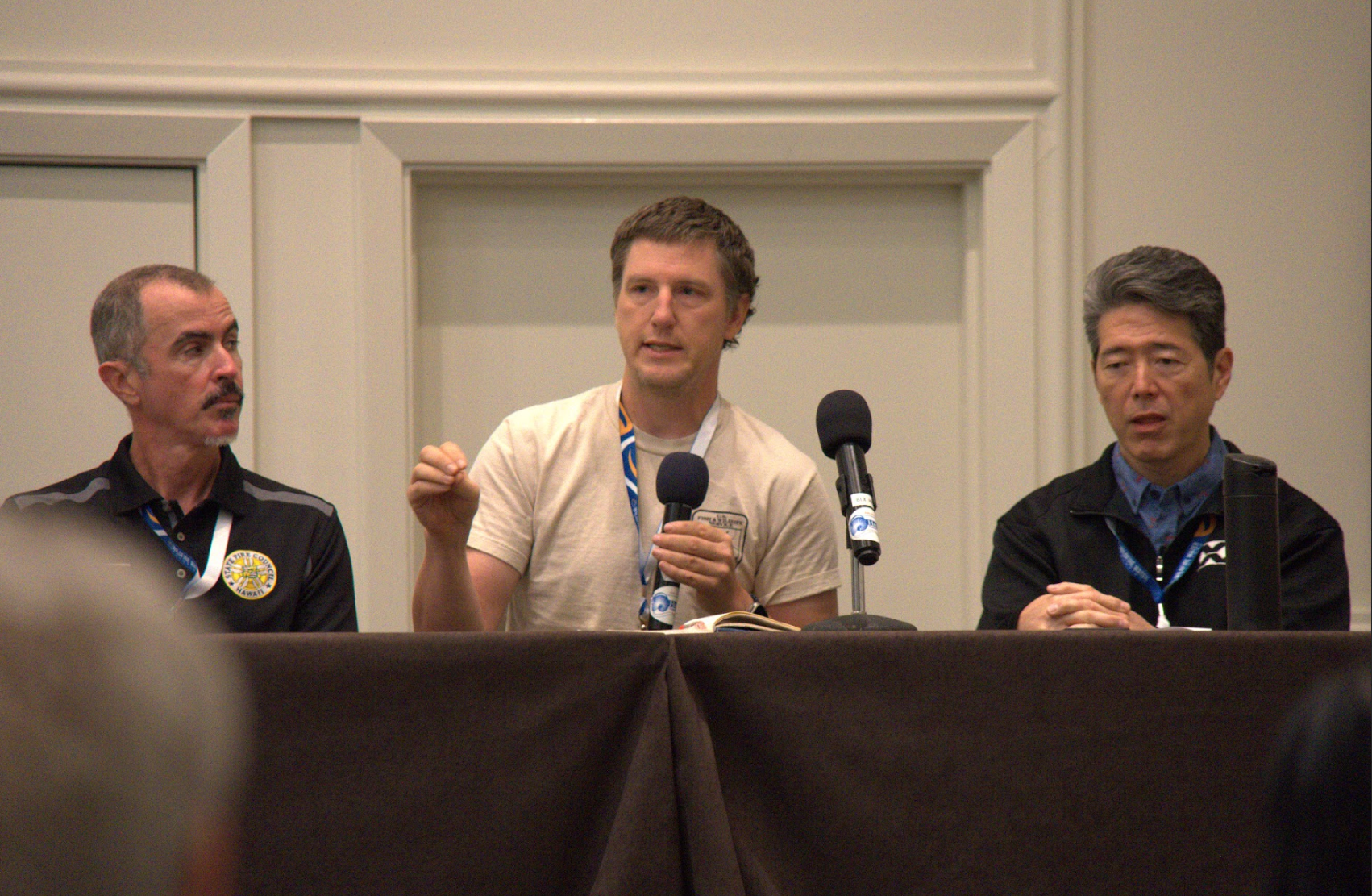 Three men sitting at a panel discussion, with one in the center speaking into microphones, the other two listening.