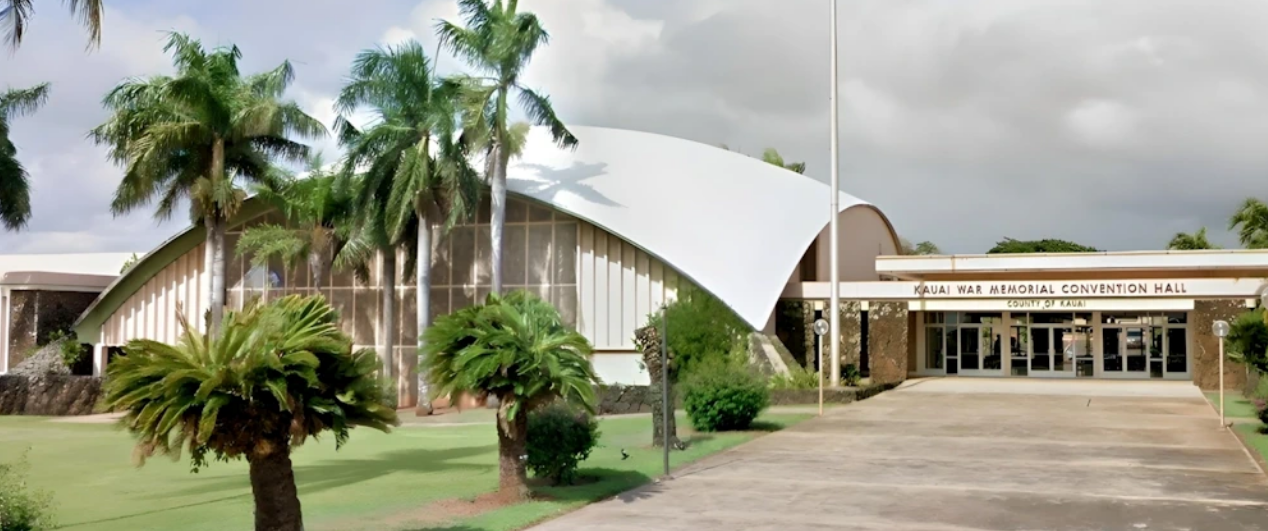 The Kauai War Memorial Convention Hall in Kauai, with palm trees, a sidewalk, and cloudy sky.