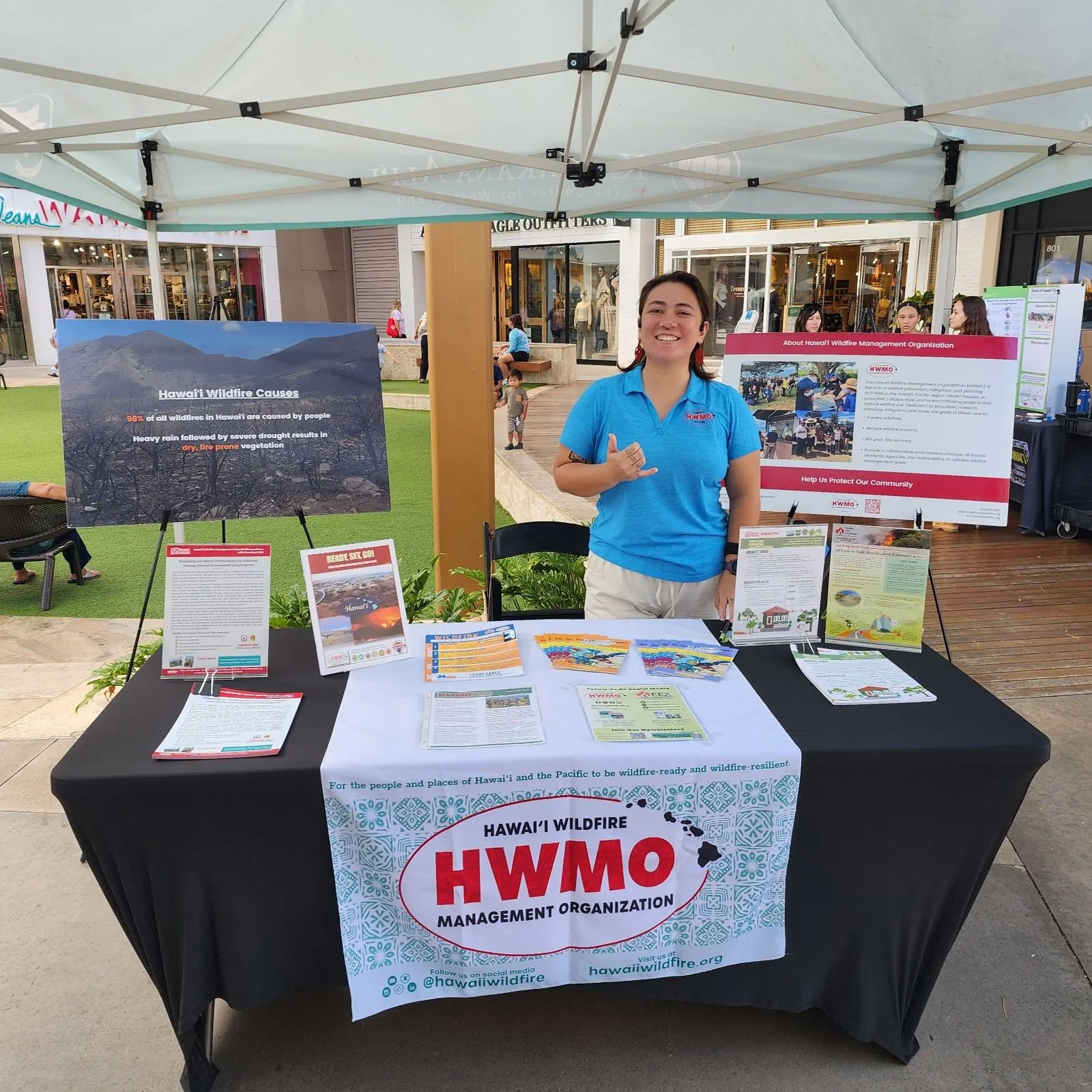 A woman standing behind a booth at an outdoor event, representing the Hawaii Wildfire Management Organization. The booth has informational posters and flyers about wildfire causes and prevention in Hawaii. The woman is smiling and wearing a blue shirt with the organization’s logo.