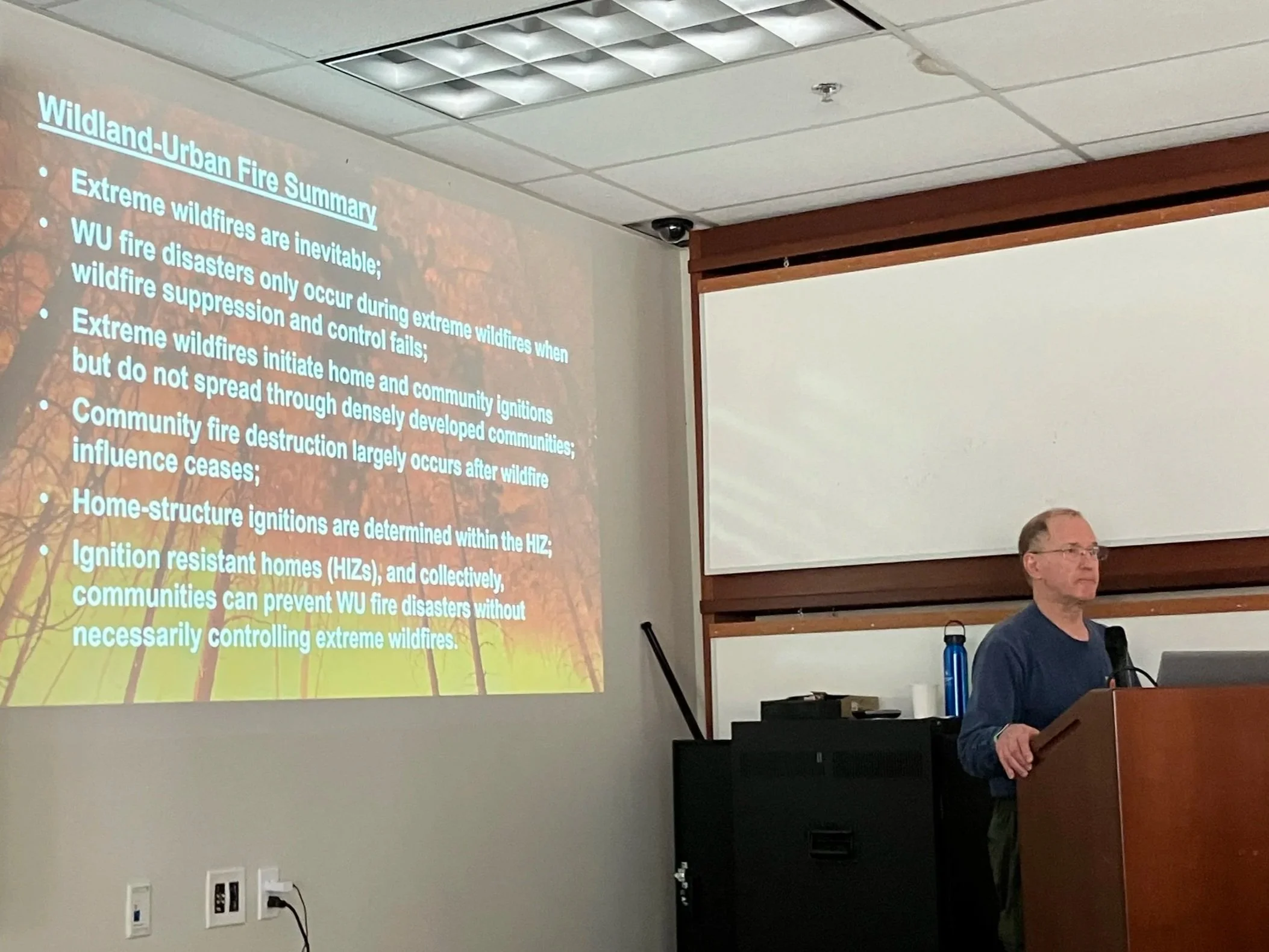 A man giving a presentation about wildfire management, standing behind a podium in a classroom. A slide on the screen is titled 'Wildland-Urban Fire Summary' and contains bullet points about wildfire prevention and community measures.