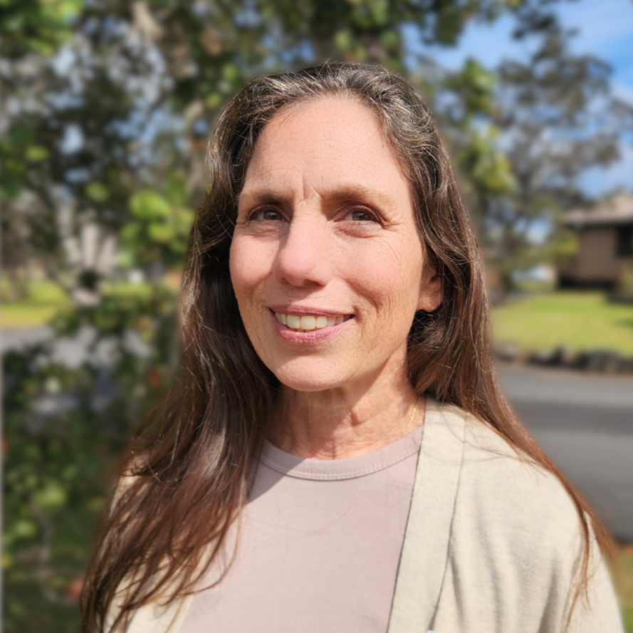 A woman smiling outdoors wearing a white cap, surrounded by green foliage.