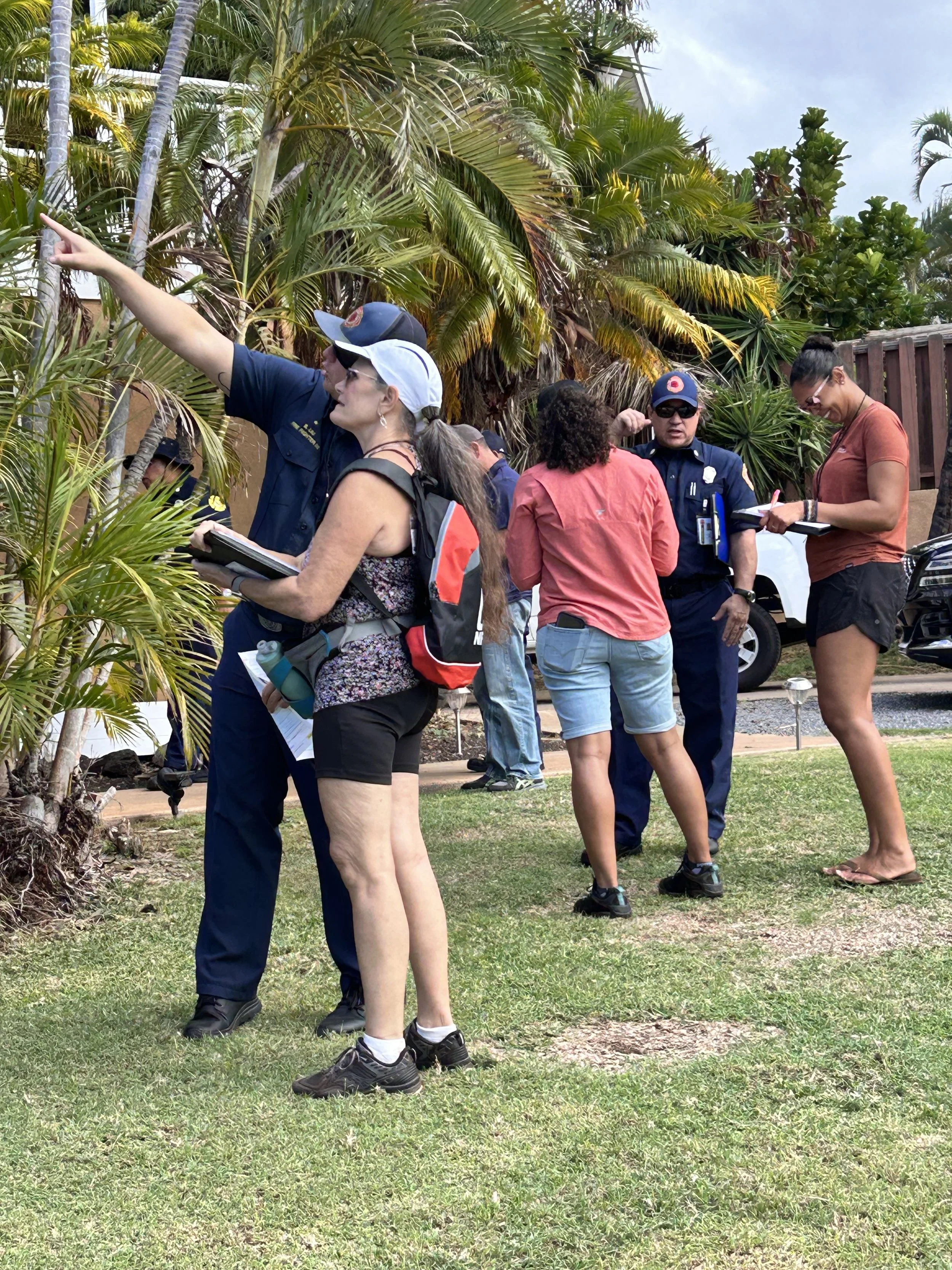 Group of people, including police officers and civilians, standing on grass near tropical plants, engaged in discussion or investigation.