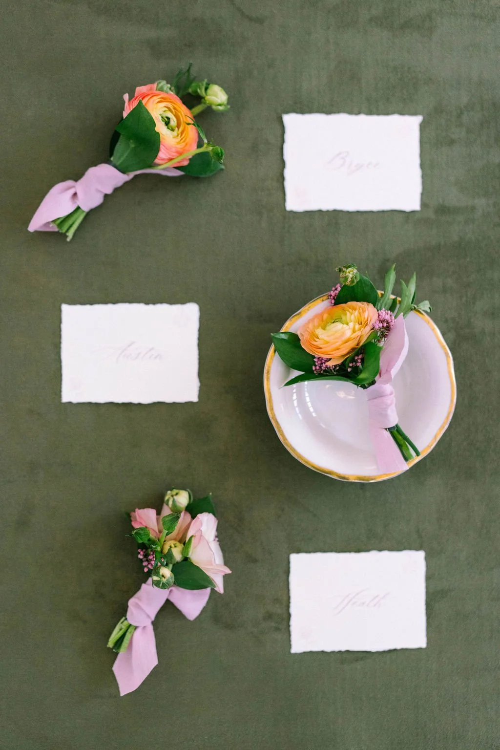 Three place cards labeled 'Bigs,' 'Matla,' and 'Maddie,' each paired with small floral boutonnieres attached with pink ribbon, arranged on a dark green table with a scalloped bowl with a floral boutonniere.