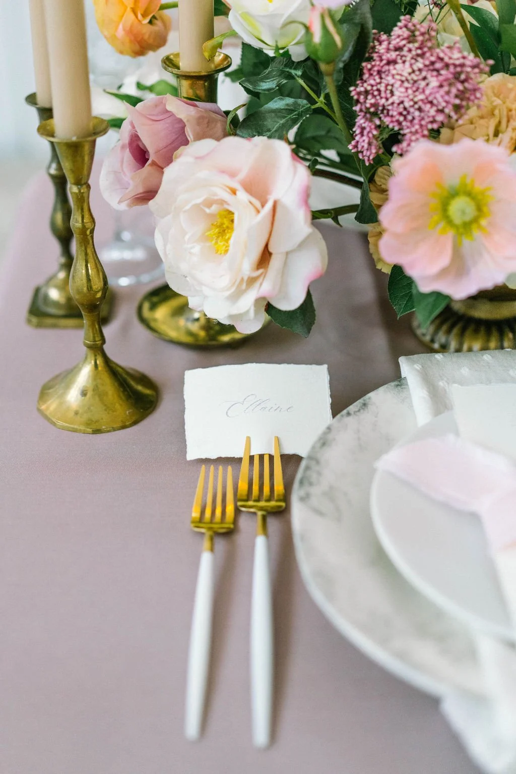 Elegant table setting with pink and cream flowers in vases, gold candlesticks, a card with the name 'Elyse', and gold and white cutlery on a light purple tablecloth.
