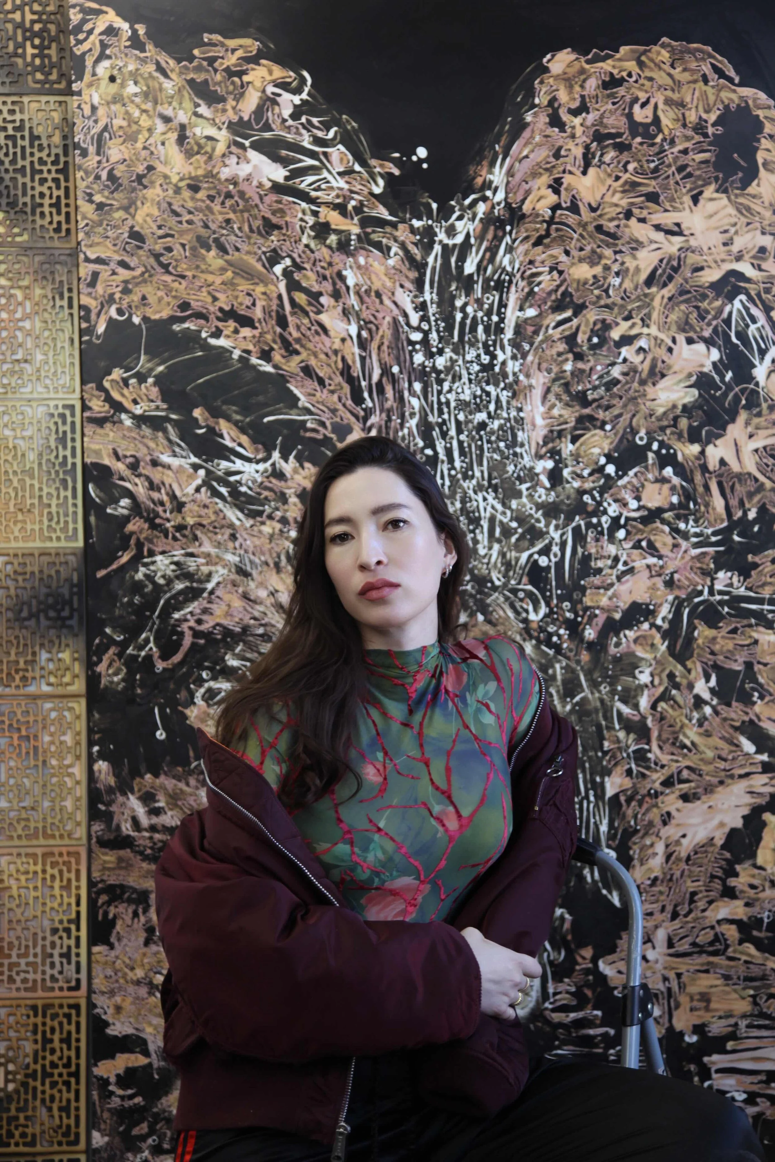 A person with long, brown hair in a mesh, red-veined dress and burgundy bomber jacket leans on a stepladder and stares directly into the camera. They sit in front of a wall work that features abstract, splatter-like brushstrokes of beige and white.