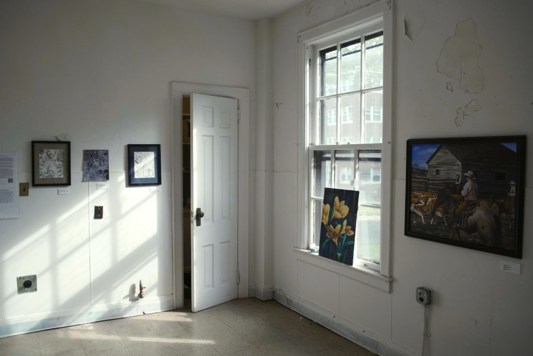 A room in a house with peeling white paint features multiple 2D works on tis walls. Light flows in from a large paned window.