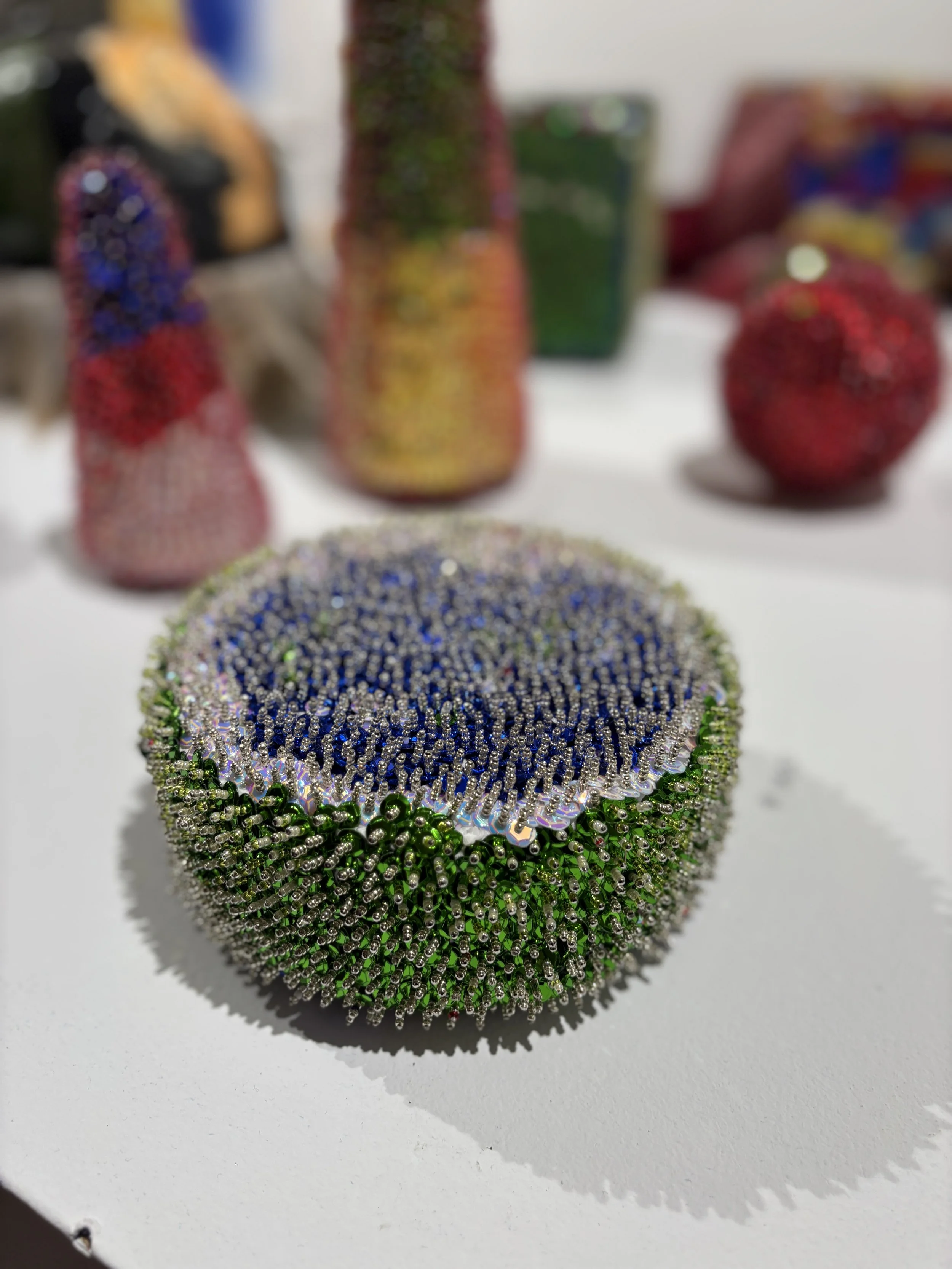Close-up of a round, spiky beaded sculpture with green sides and a blue center, surrounded by other blurred beadwork.