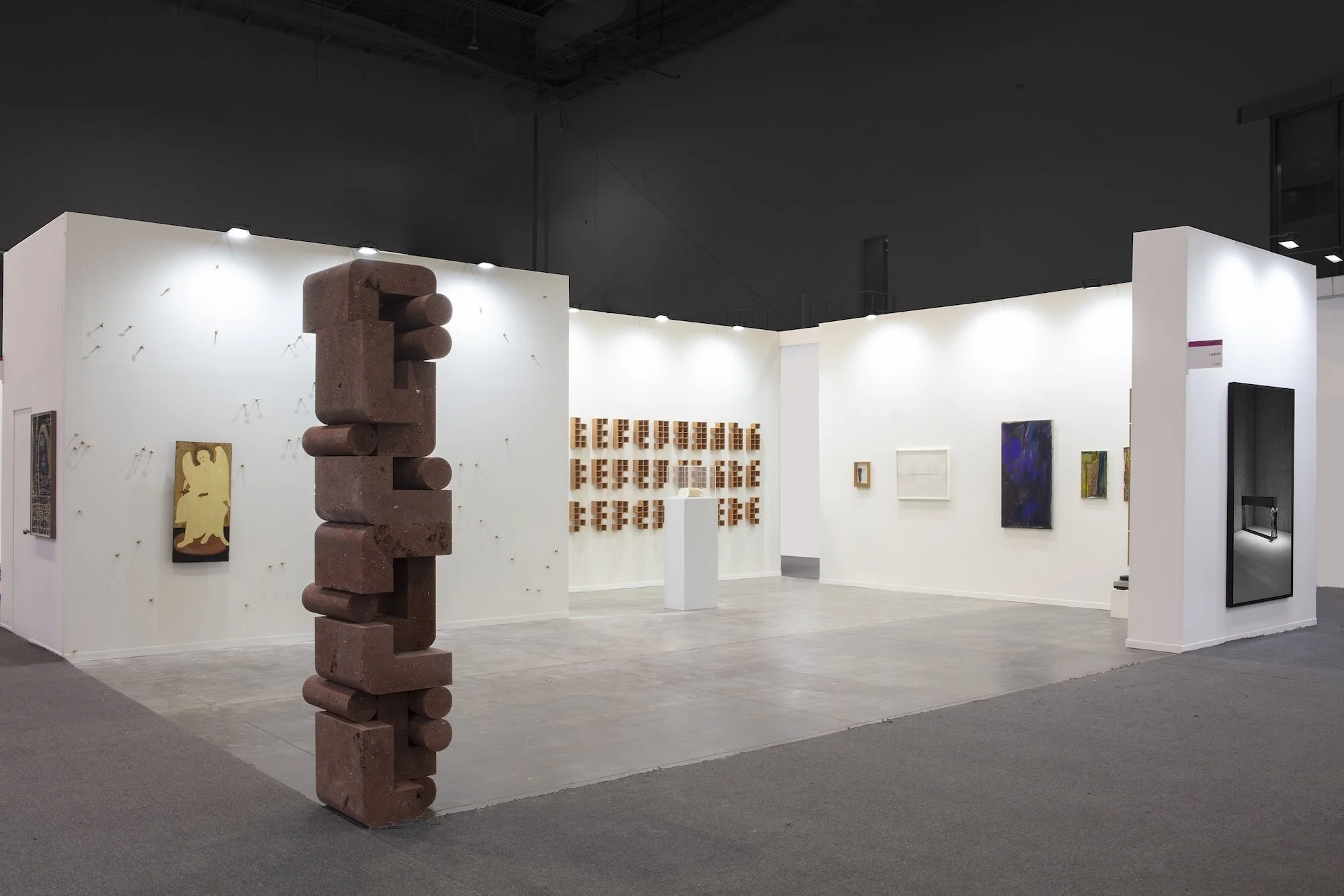 Art fair booth view featuring a tall stacked sculptural column, small wall-mounted works, and minimalist plinths under bright exhibition lighting.