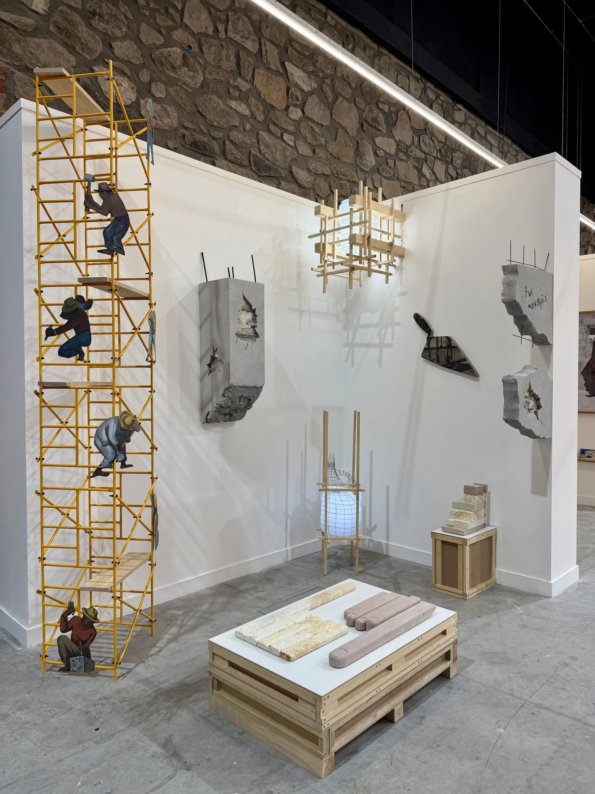 Installation view of mixed-media sculptures, including a yellow scaffold with painted figures, concrete fragments, and wooden structures arranged in a white-walled gallery.