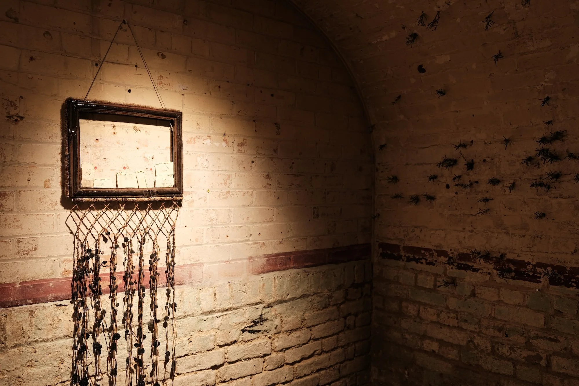 Framed mirror with hanging cords and beads mounted on a brick wall, surrounded by scattered black insect-like forms.