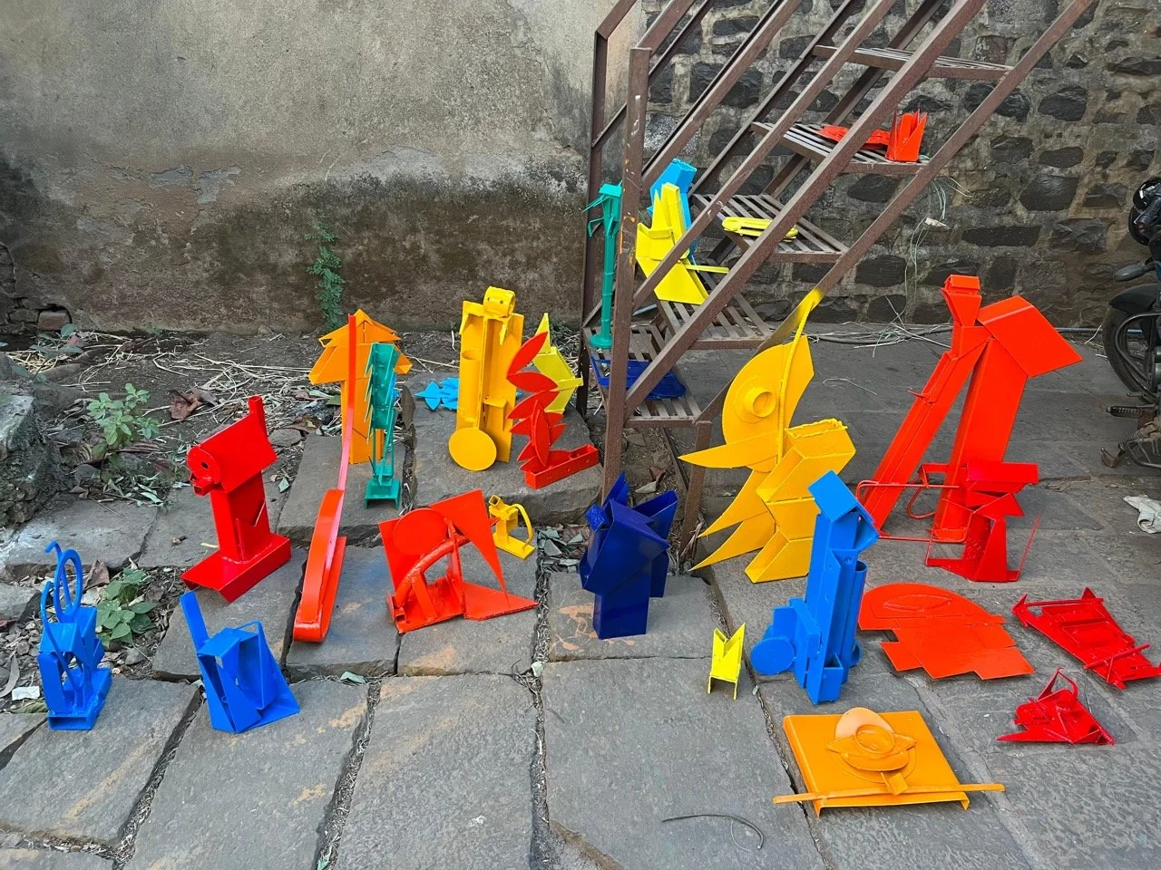 Colorful abstract metal sculptures arranged on stone pavement beside an outdoor staircase.