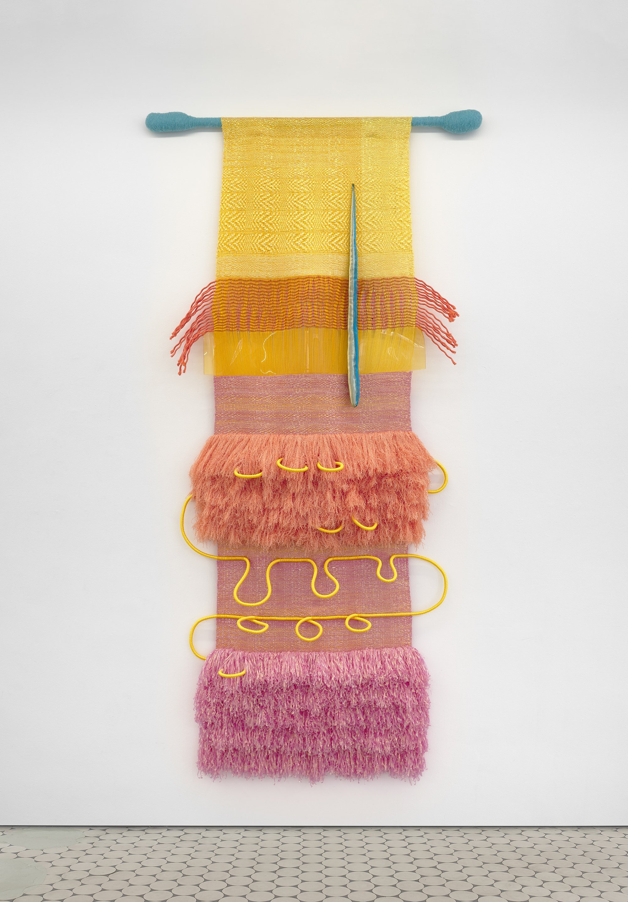 A fiber work mounted to a white wall is made from layers of yellow, pink, and orange fiber, some tufted, some thick and wound. The piece hangs from a rod wrapped with blue thread.