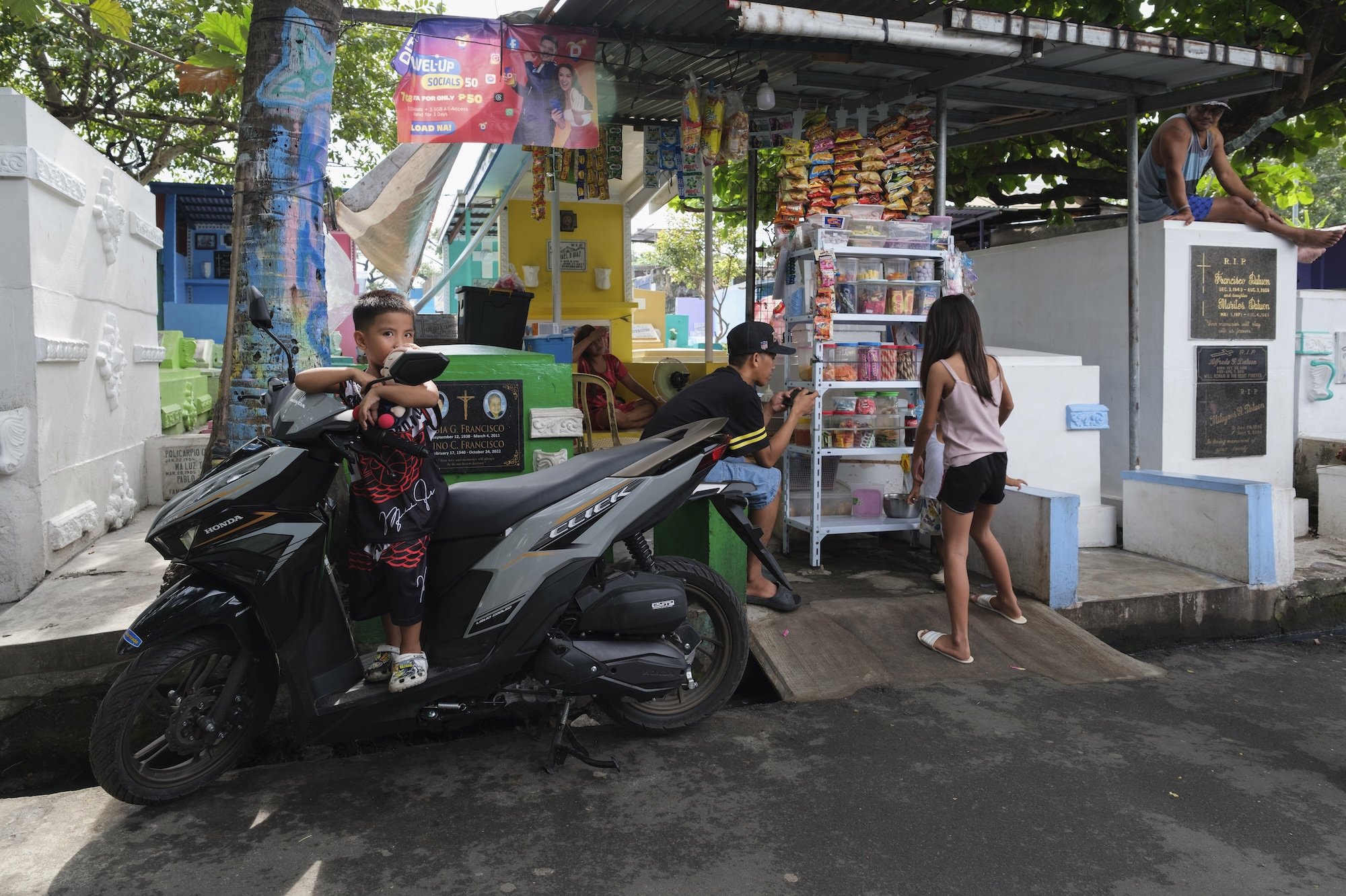 Street snack stand set up beside cemetery tombs, where children and adults gather near a parked motorbike and shelves of packaged food.
