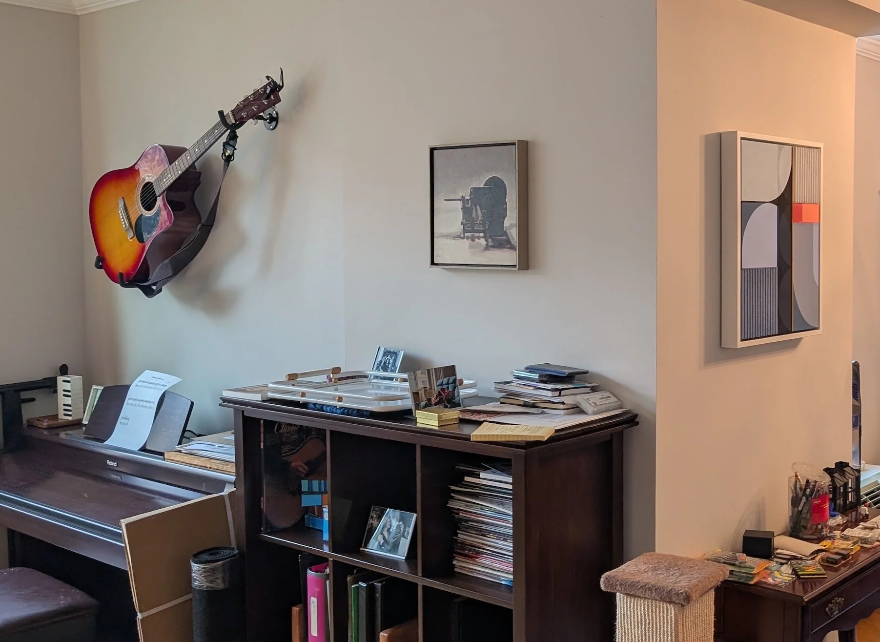 A living room corner with a wall-mounted acoustic guitar, framed artworks, a keyboard, and shelves filled with records and books.
