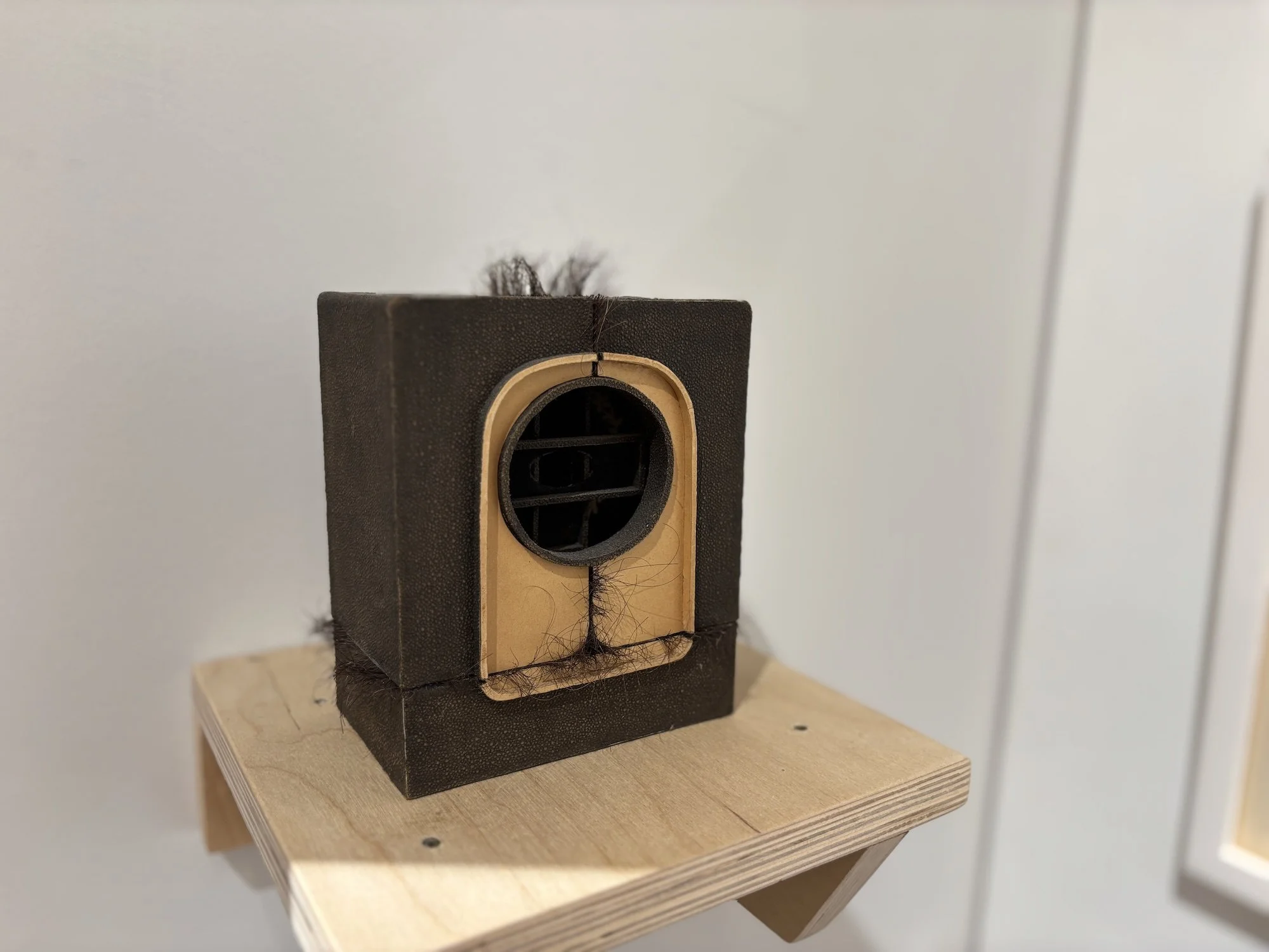 Small sculptural object resembling a vintage radio, wrapped in dark material with strands of hair emerging from its seams.