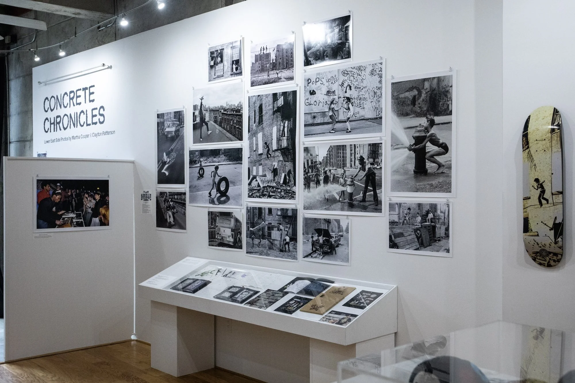 Gallery installation with black-and-white photographs displayed salon-style above a vitrine of archival materials.