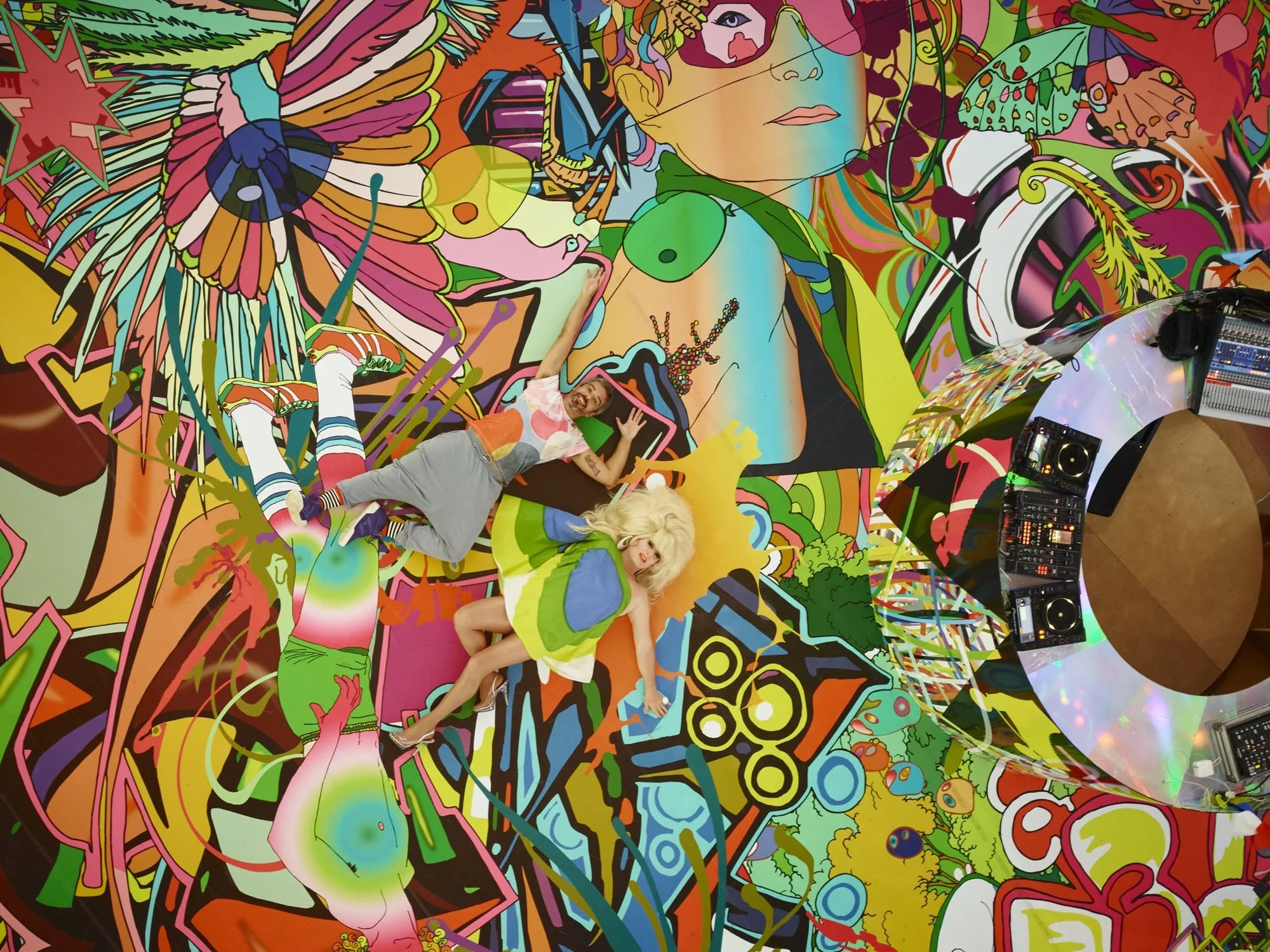 On a floor made up of colorful illustrations of people, flowers, and other abstract patterns, two people lie on the floor and pose for an aerial shot. To the left, a holographic circular DJ booth stands.