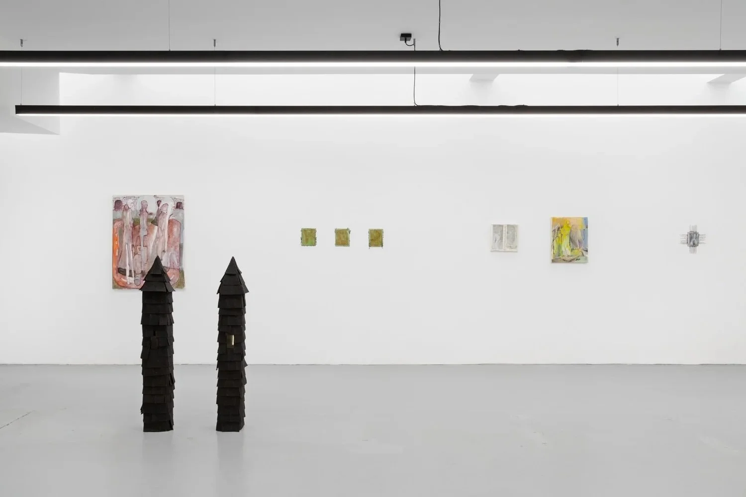 In a white gallery room, numerous small paintings in soft colors sparsely populate the walls. On the grey floor, two freestanding sculptures appear as dark turrets.