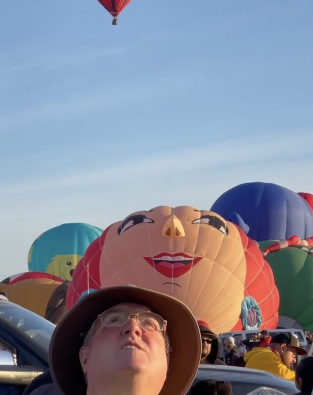 Hot air balloons shaped like cartoon faces rise over a crowd at a balloon festival.