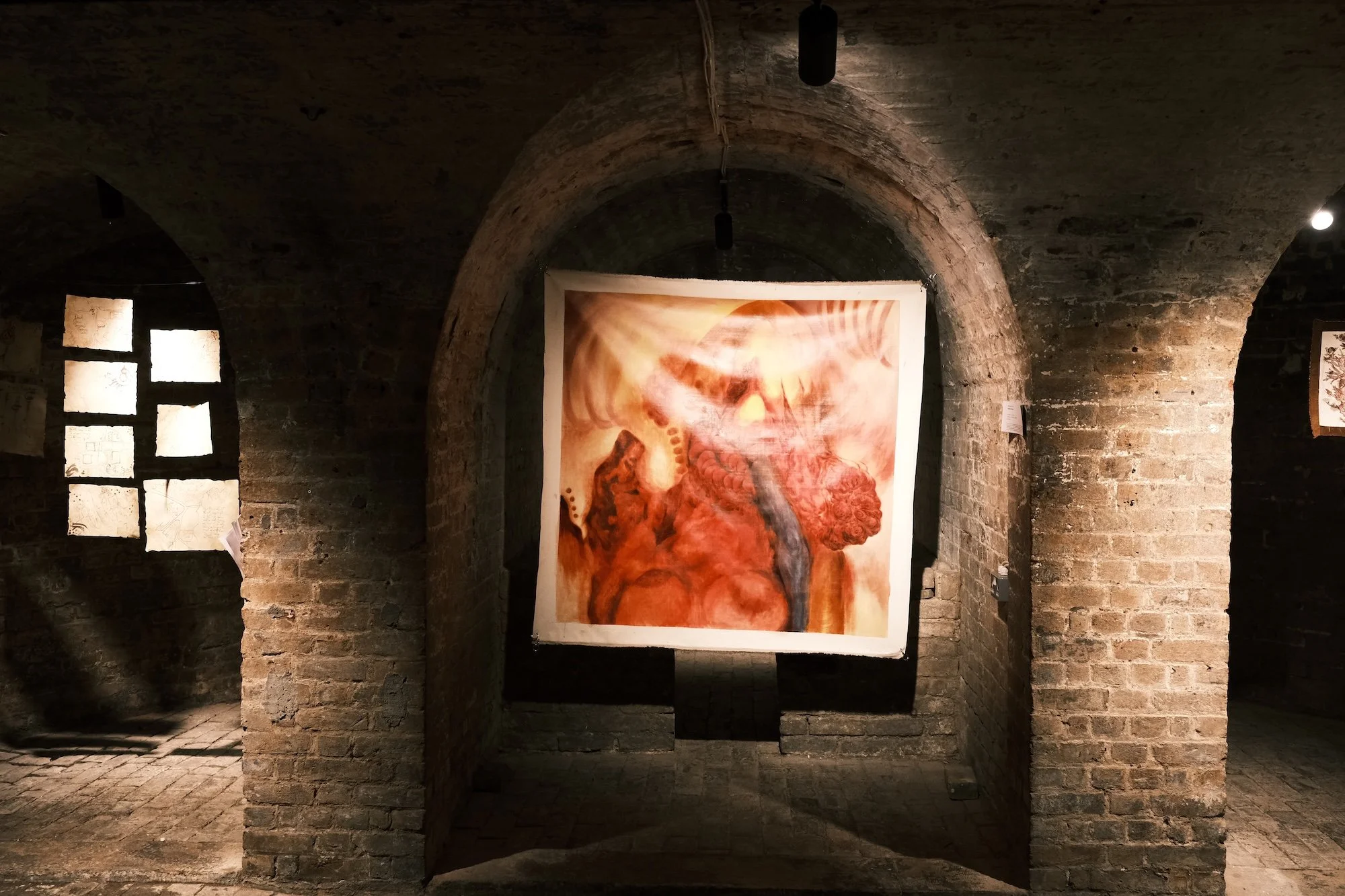 Large semi-transparent painting in fleshy reds and browns hangs between brick arches in a vaulted underground space.