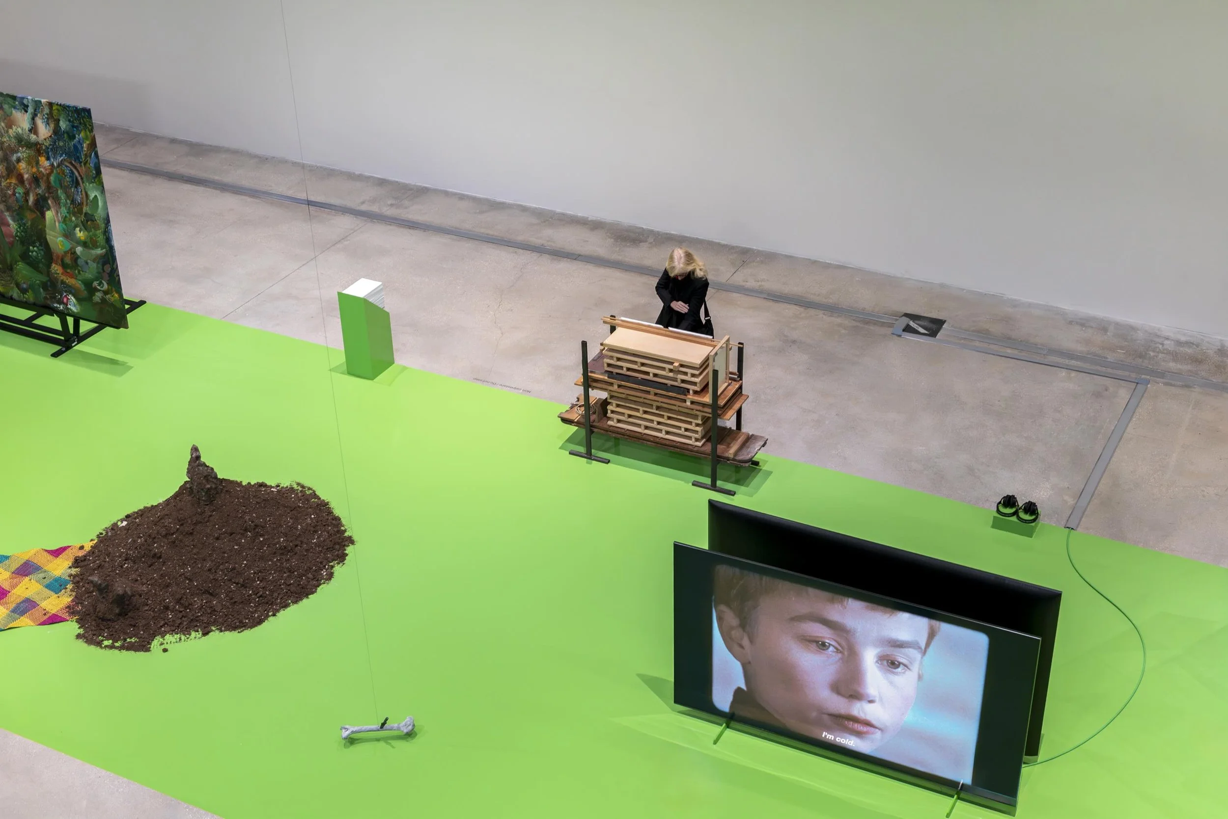 A large white room with concrete floor is partly covered with a green  surfacing.  On the green surfacing are multiple artworks of different mediums: there is a TV, a pile of dirt, and other assorted sculptures.