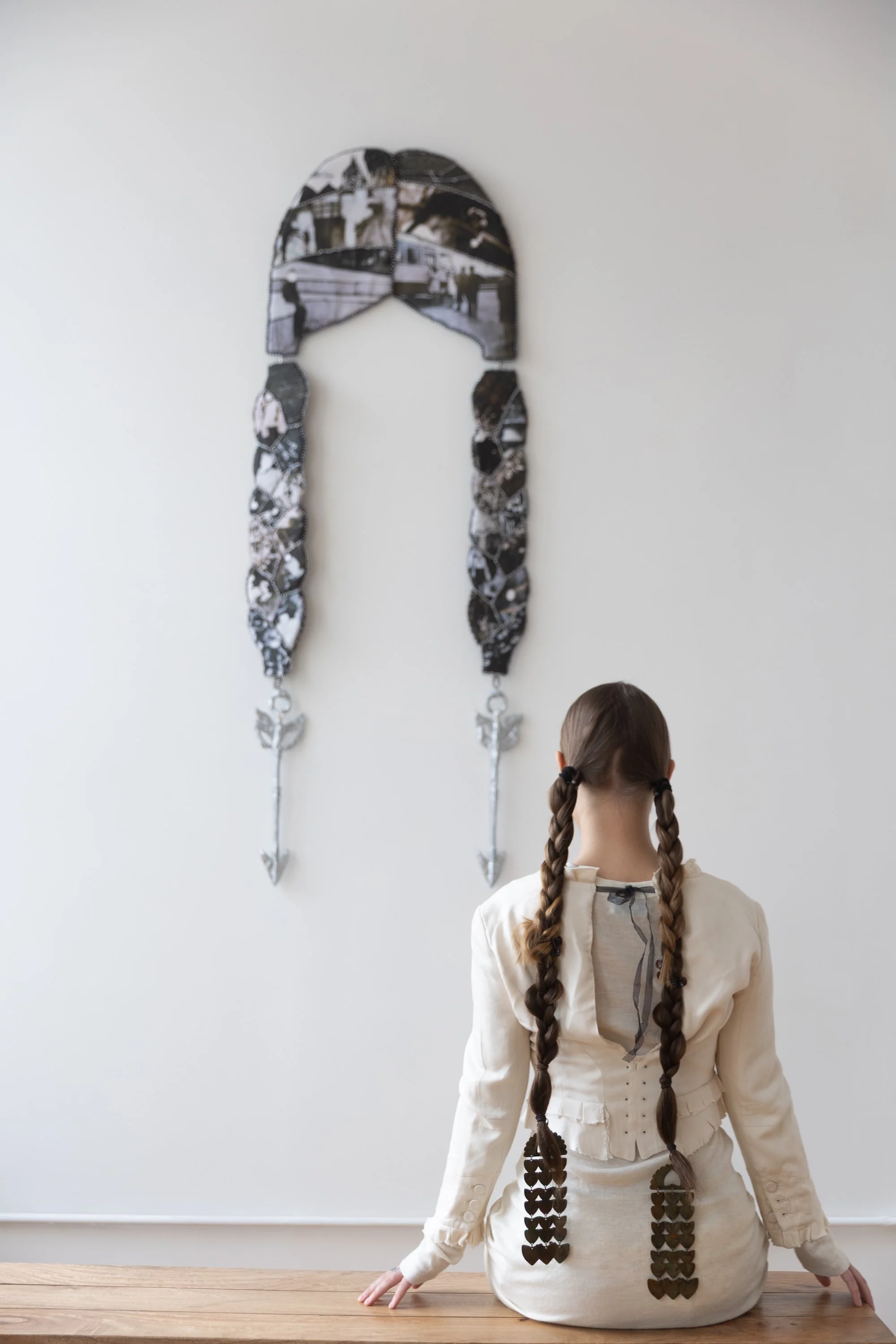 Woman with long braided pigtails sits facing a wall-mounted artwork shaped like braids made from stitched black-and-white photographs with silver arrow pendants.