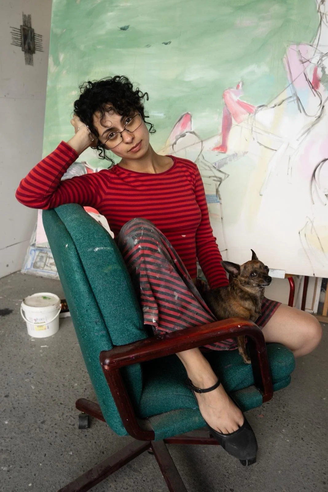 In a room littered with paint and a large canvas, a person in a red striped dress sits diagonally in an upholstered wooden rolling chair. Her head rests on her hand and a small brown dog sits in her lap.