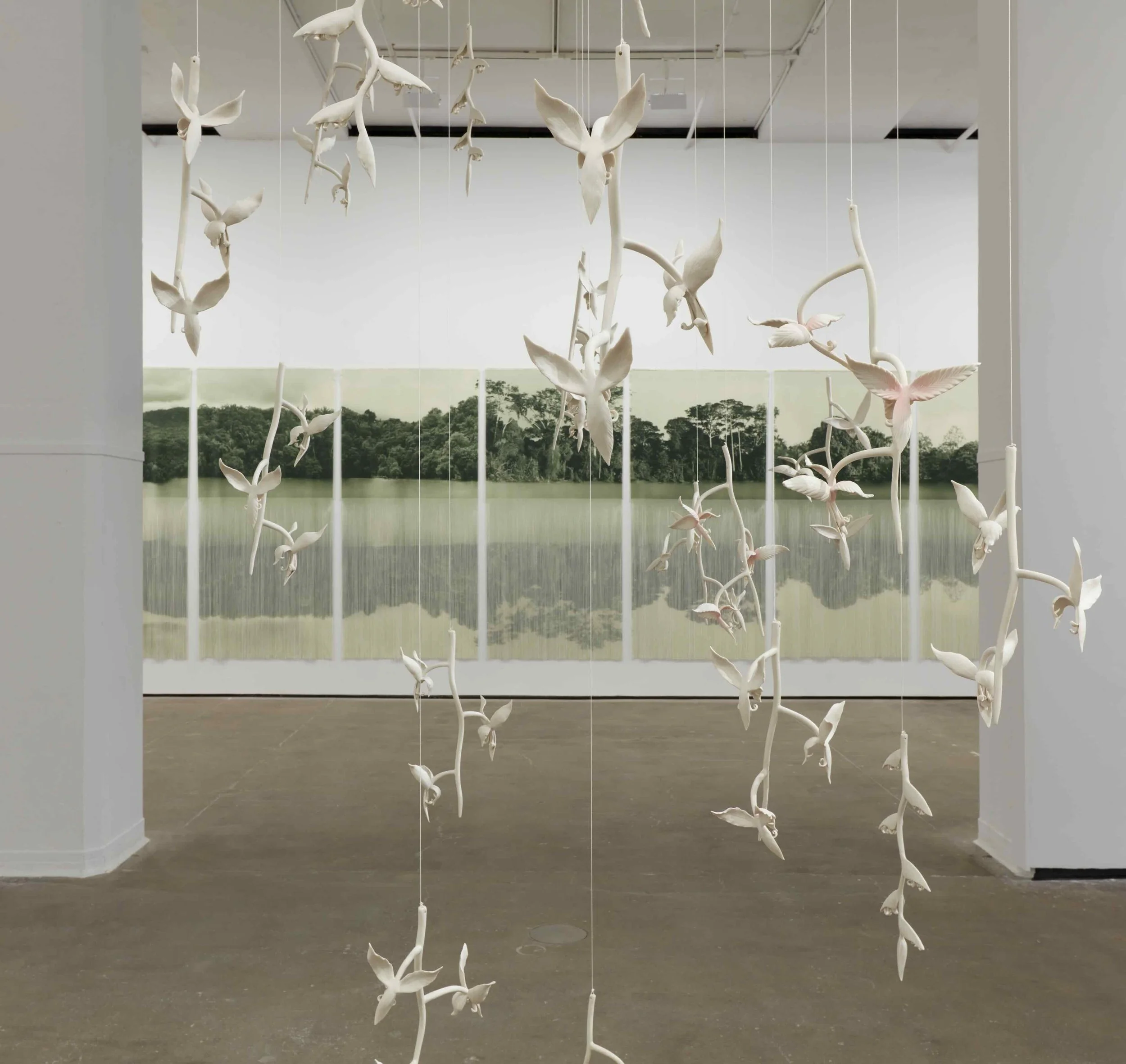 A multi-room gallery has a large opening that reveals a large textile work in the next room. In the foreground room is a hanging sculpture comprised of invisible wire and white porcelain flowers and plants.