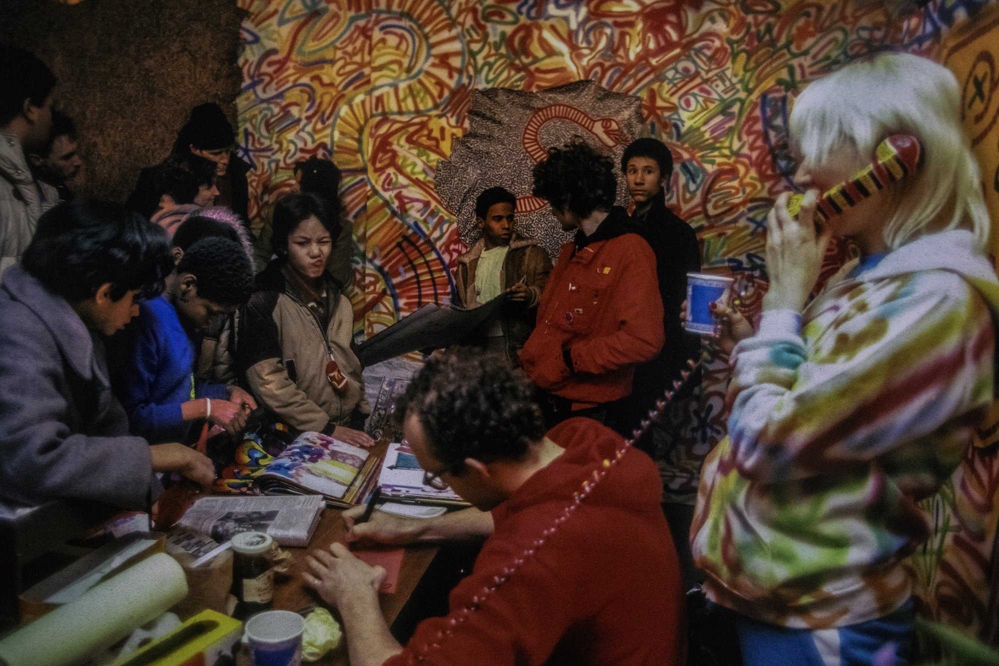 Group gathered around a table covered in zines and papers in front of a brightly painted mural.