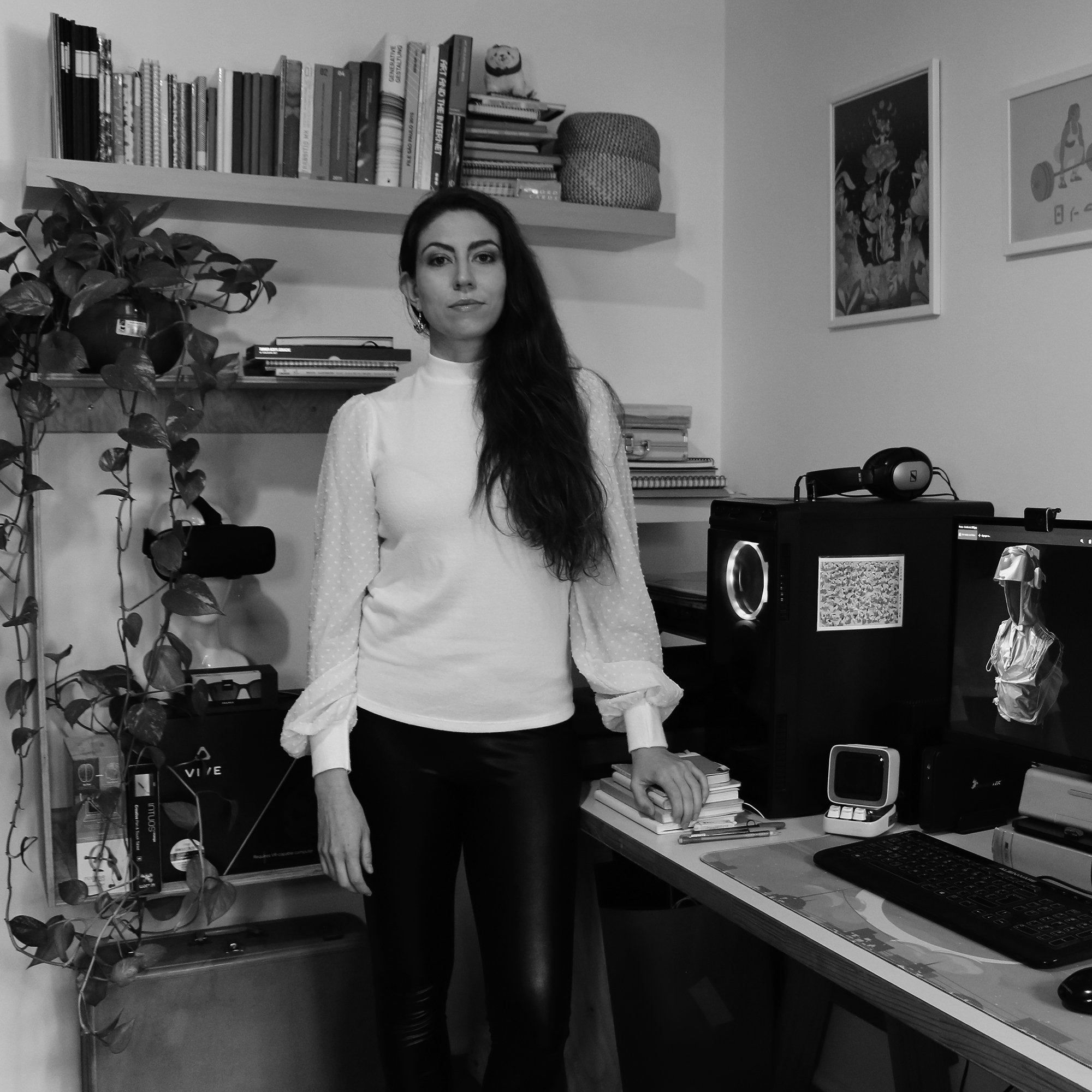 Woman standing in a home office beside a computer setup and shelves of books and plants, wearing a white blouse and black pants.