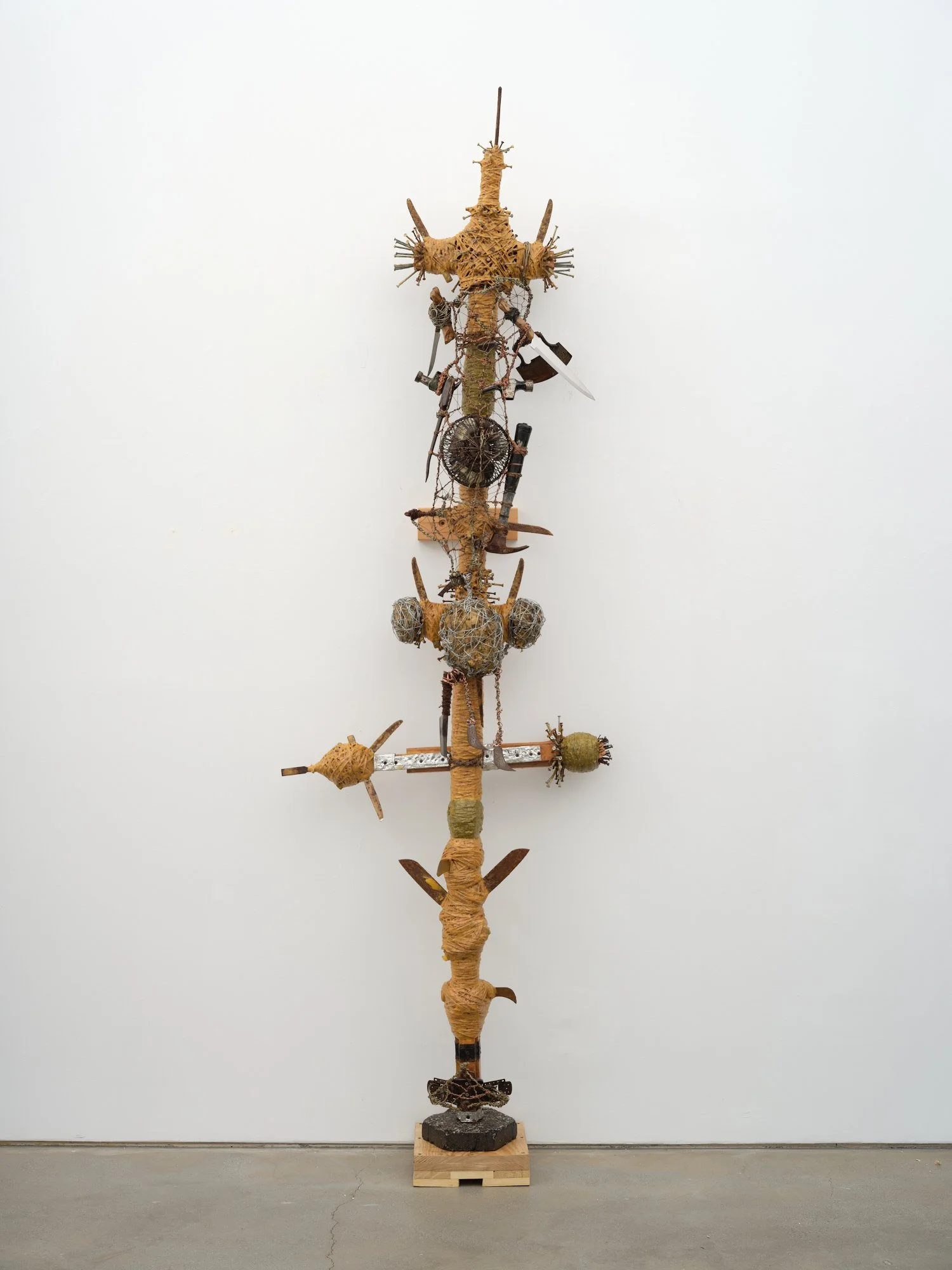 A tall, totem-like assemblage of wrapped wood, wire, tools, and found objects stacked vertically on a small wooden base.