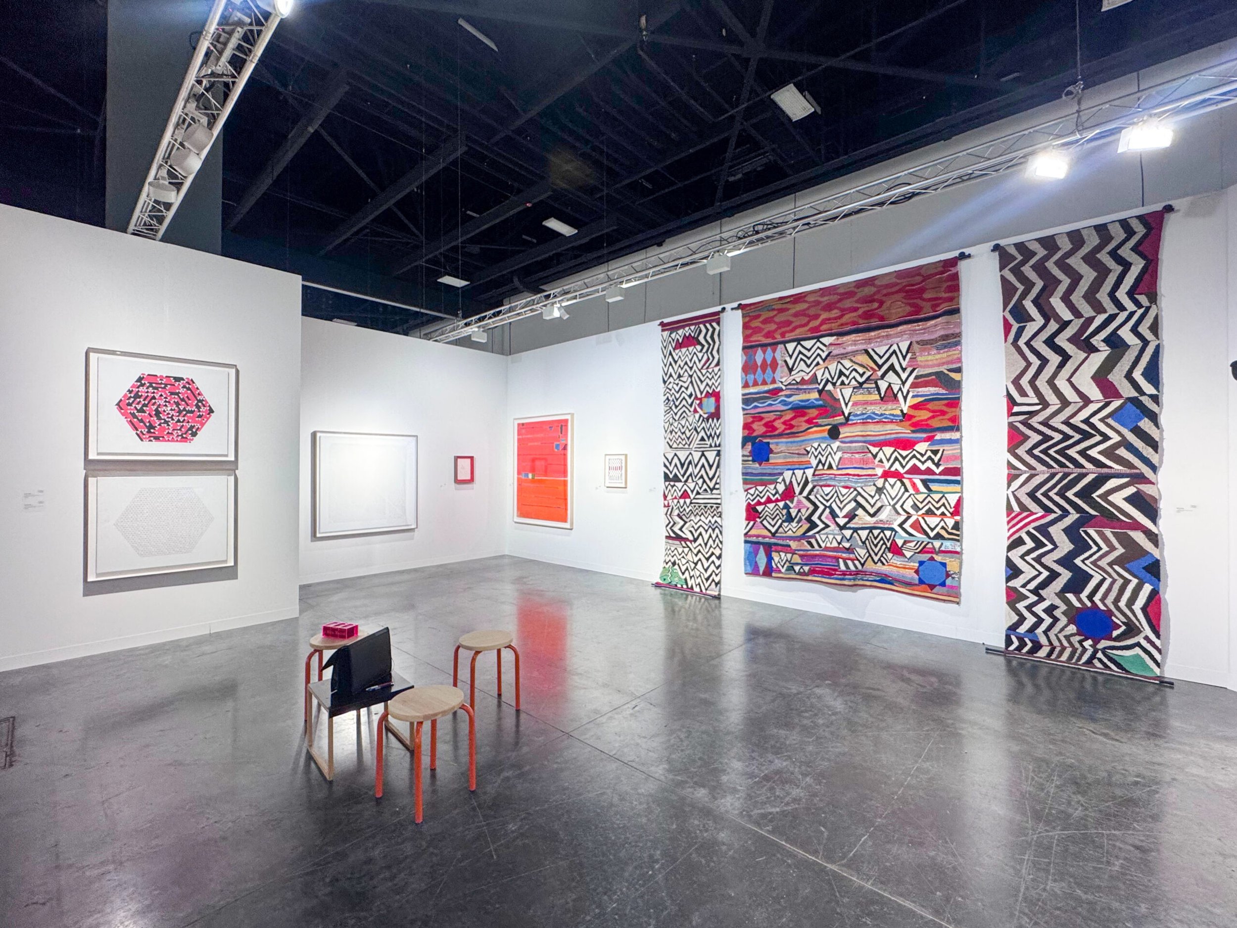 Art fair booth with framed geometric works and three large, colorful patterned textiles hanging on the right wall.