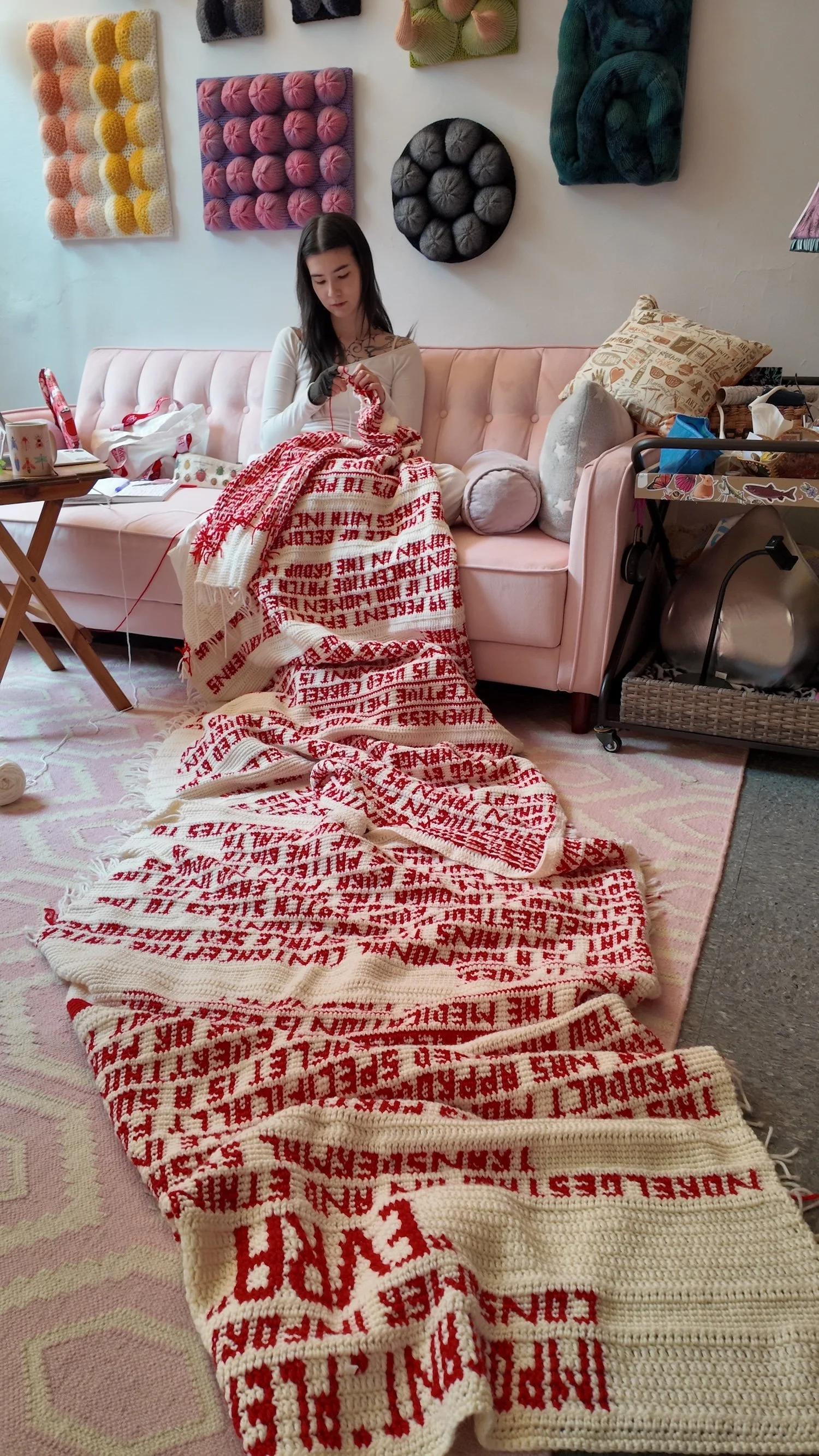 Artist knitting a long red-and-white text-based crochet tapestry draped across a pink sofa and rug.