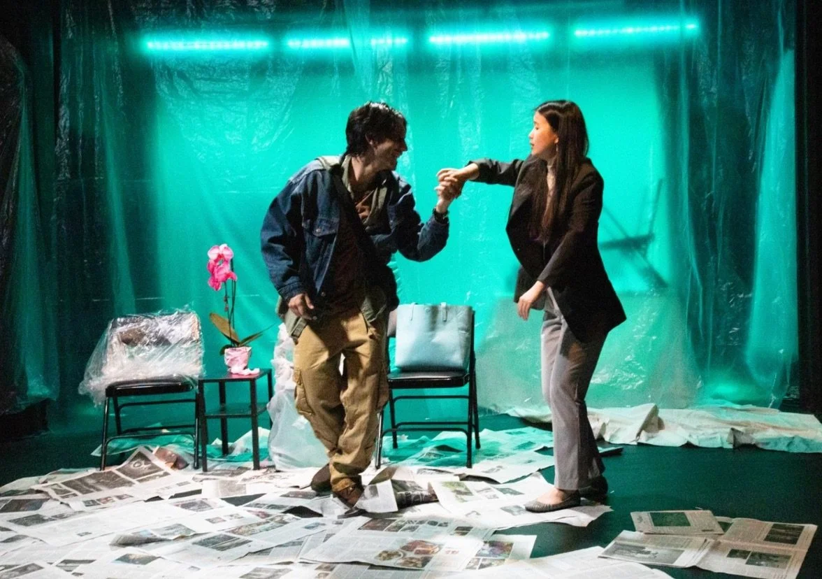 Two actors perform an intense scene on a stage covered in newspaper, with plastic-covered chairs, a pink orchid, and teal lighting creating a surreal, gritty atmosphere.