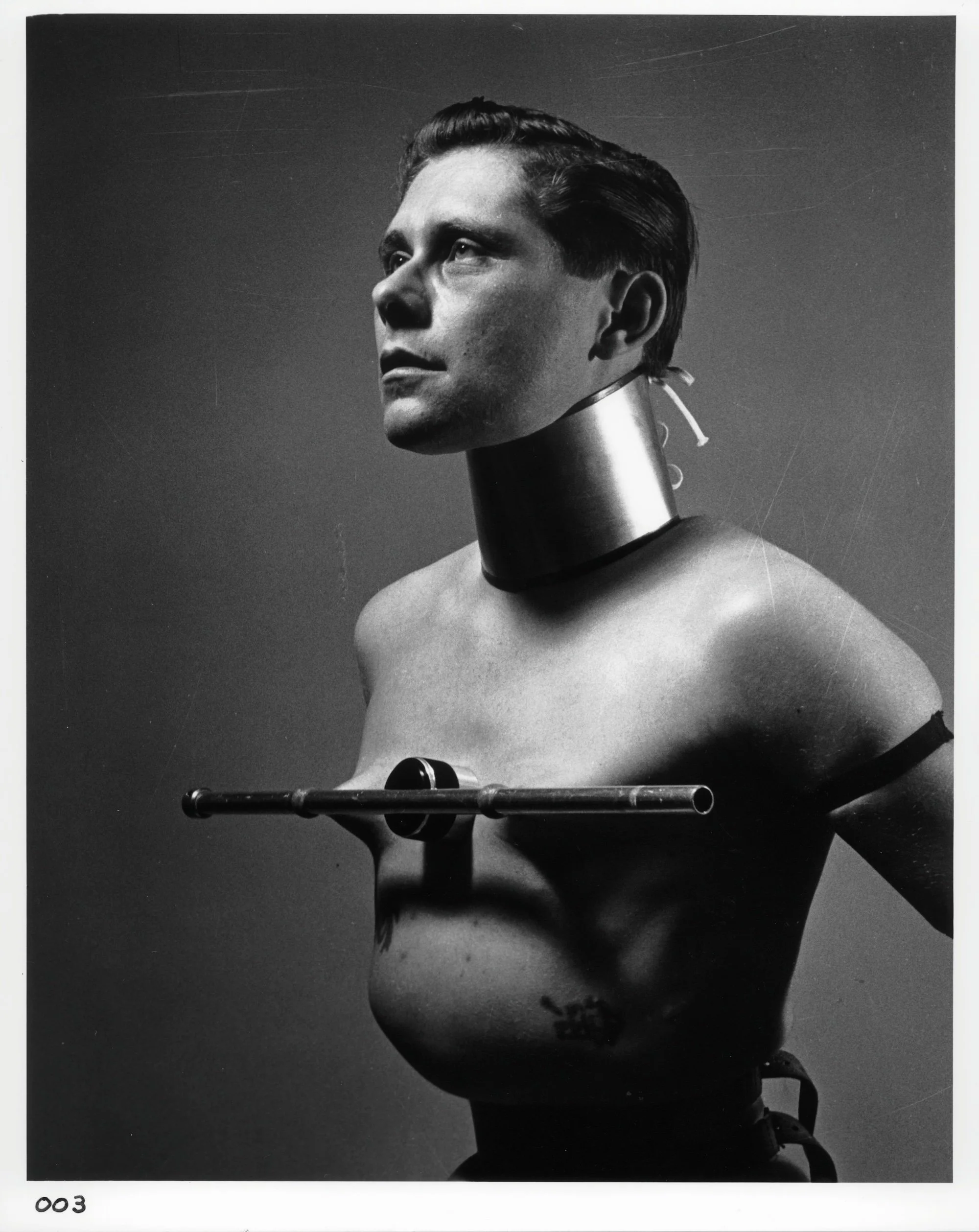 Black-and-white portrait of a bare-chested figure wearing a rigid metal collar and chest apparatus.