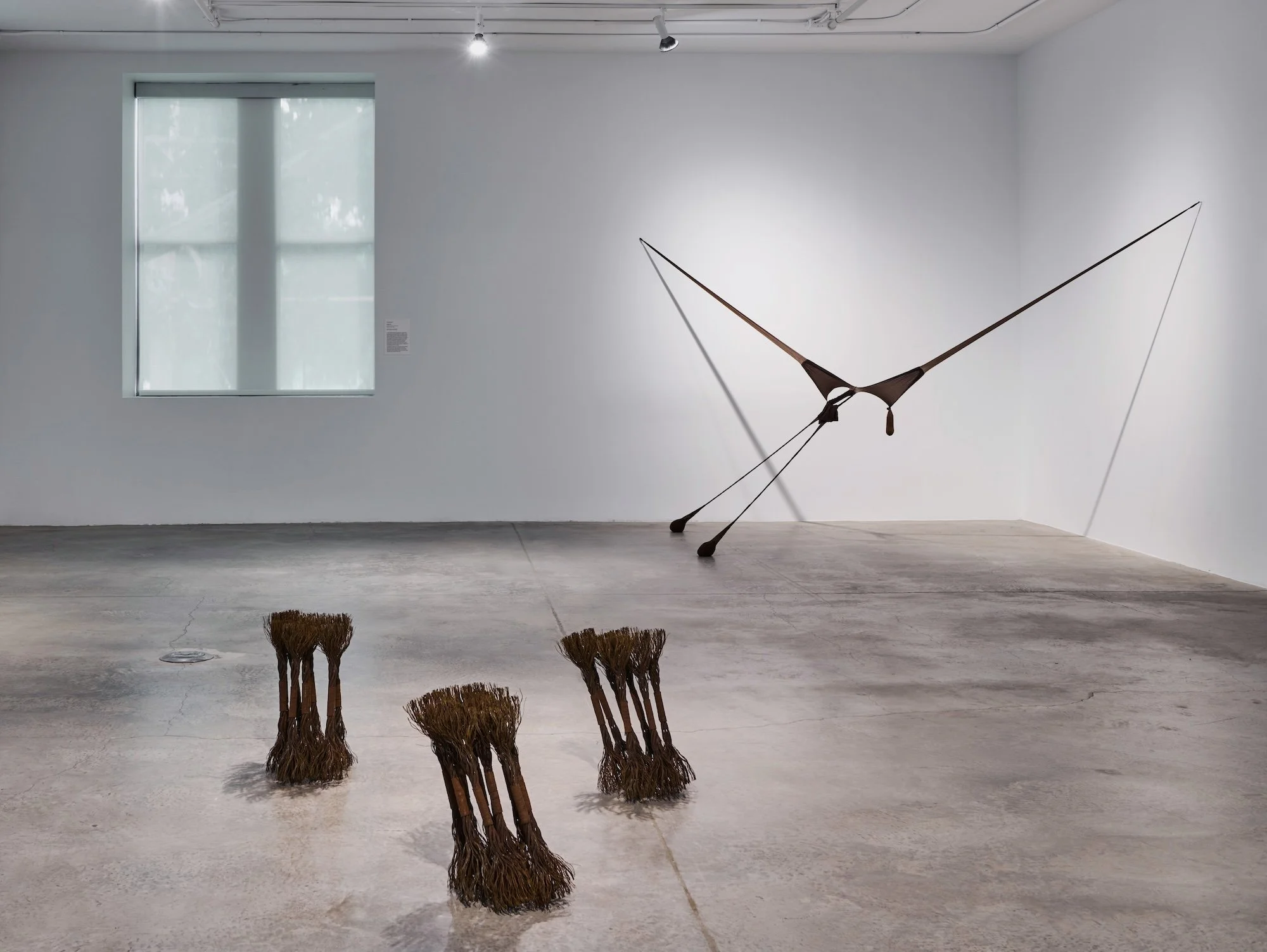 A minimalist gallery space featuring a suspended, wing-like stocking sculpture, with three small bundled brush-like forms placed on the concrete floor.