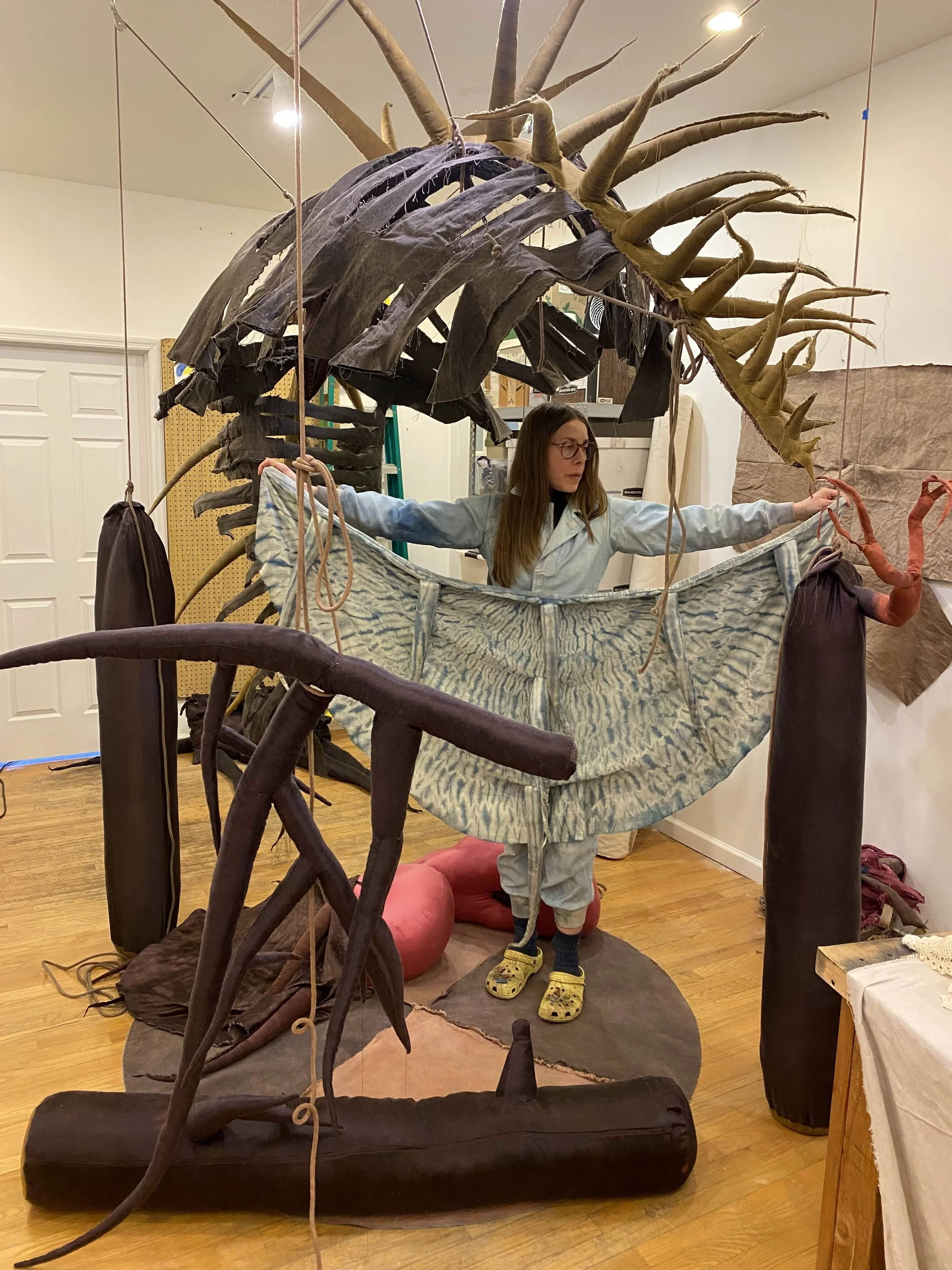 A person in coveralls stands in a room populated and hing with different fabric sculptures that take on spindly, organic forms. The person holds up a fabric sculpture in the shape of a semicircle with tendril-like attachments, tie-dyed blue and white
