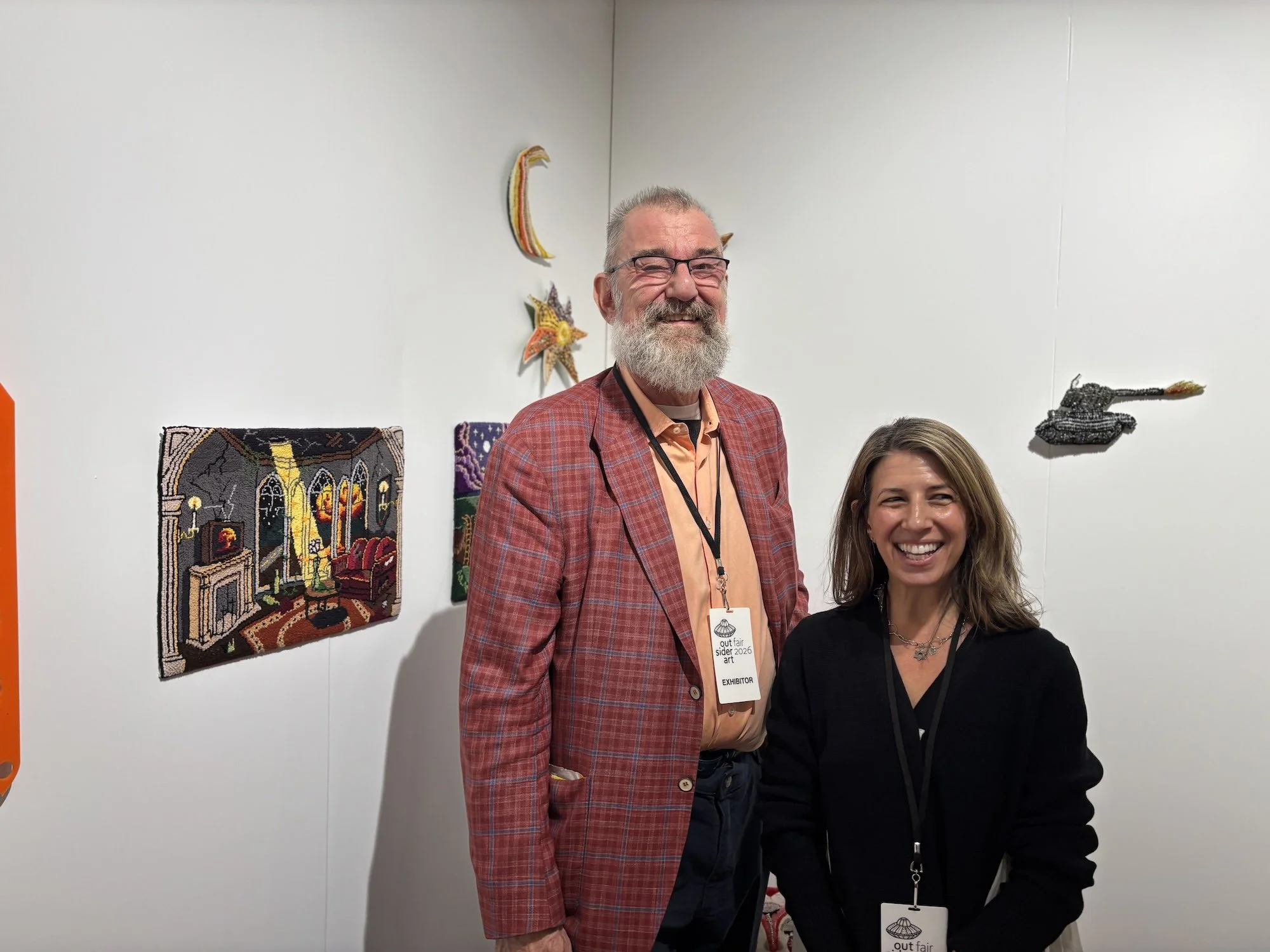 Two smiling exhibitors stand in a booth with small textile artworks displayed on a white wall behind them.