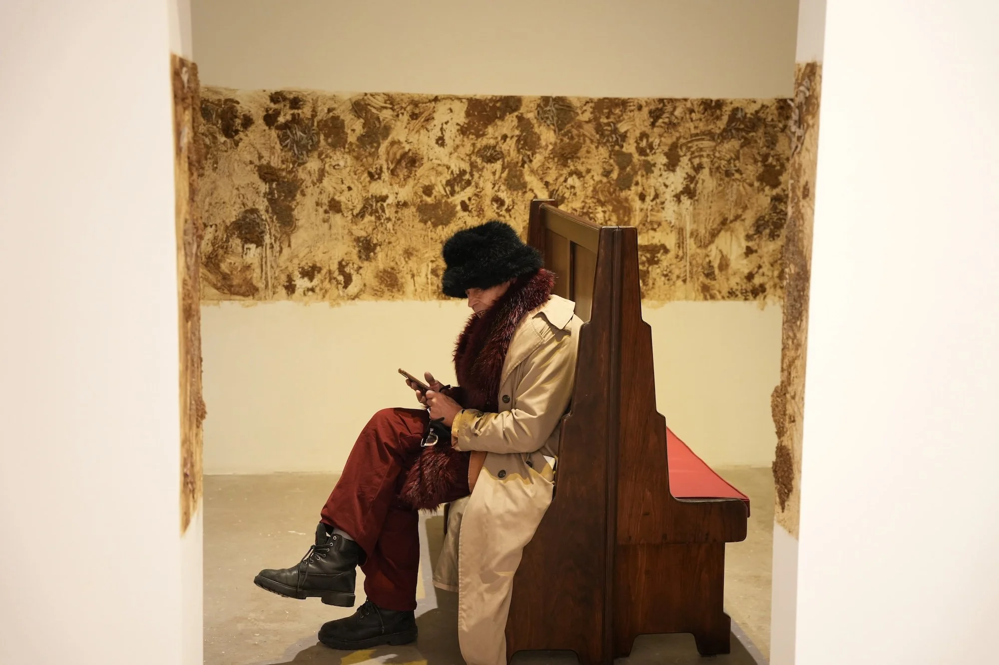 A person in a fur hat and trench coat sits on a wooden bench, looking at their phone, framed by textured brown wall pieces.