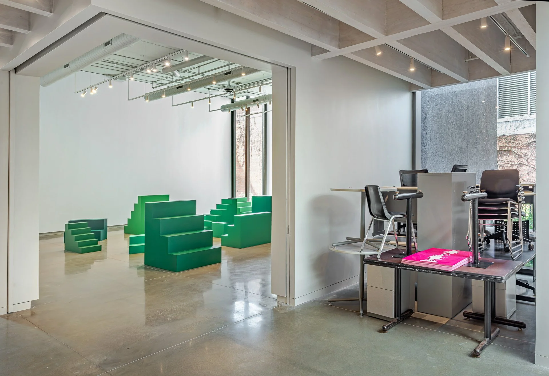 Two white gallery rooms connected by a large entryway host two installations. The left back room is populated with small and medium green staircases. The right front room is filled with classroom tables, desks, and chairs stacked on each other.