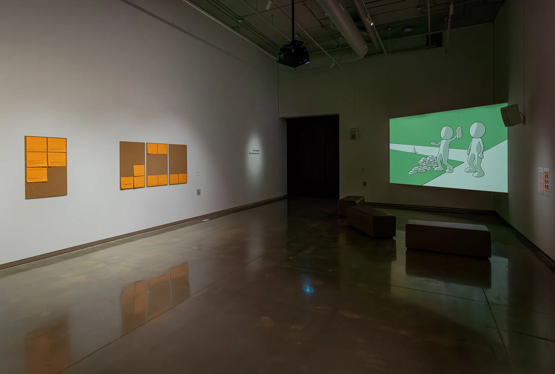 A large, dimly-lit white gallery room hosts a wall work on one wall, comprised of bulletin boards with orange slips of paper. On the far wall, a video work of illustrated white figures in a green background is projected.