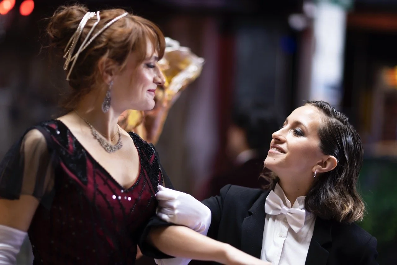 Two women in period costume — one in a beaded burgundy gown, one in a tuxedo — dancing and smiling at each other.