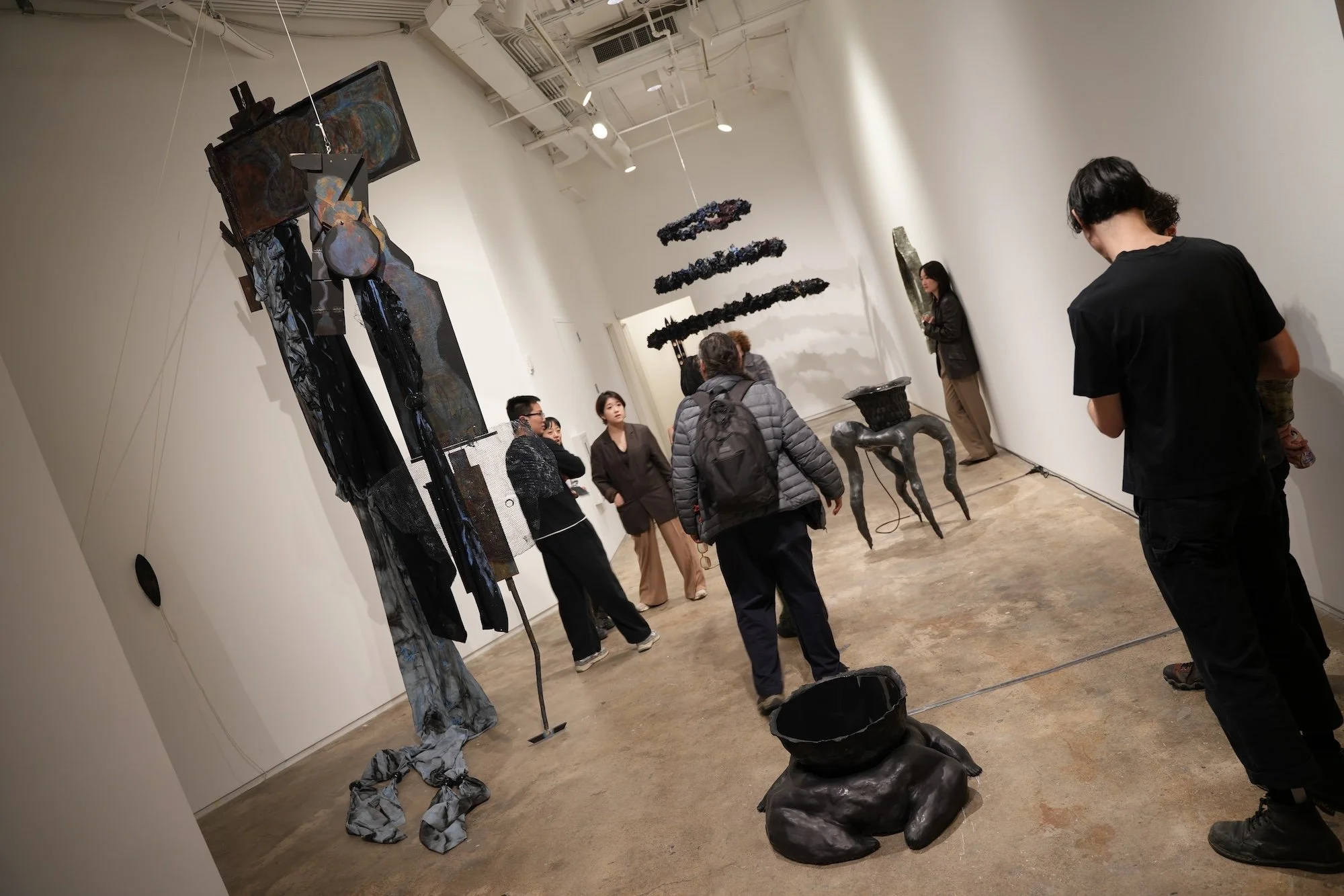 Gallery visitors walk among large, dark abstract sculptures and wall-mounted works in a white exhibition space.