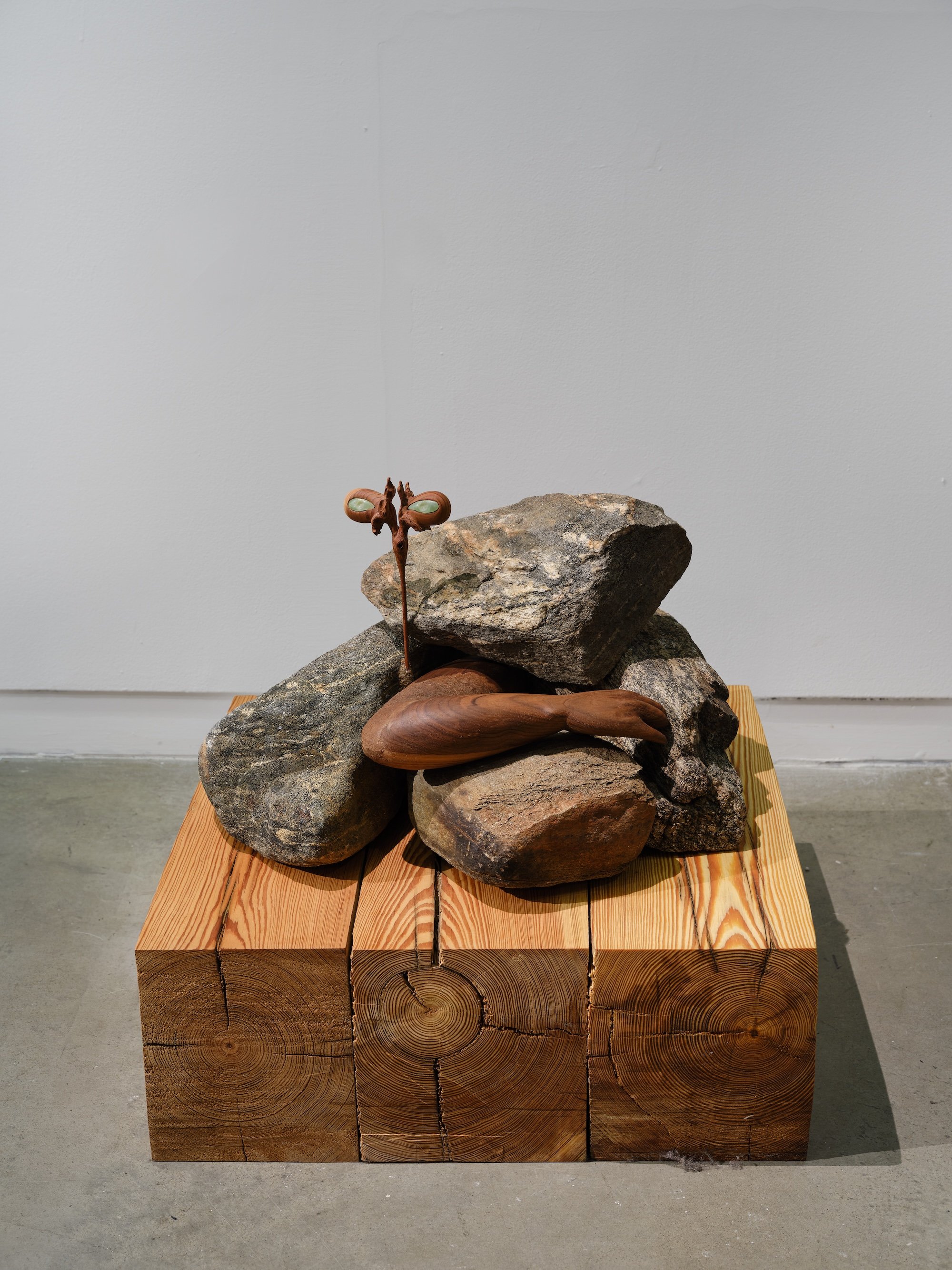 Sculptural arrangement of stacked rocks and carved wood on a wooden block base, featuring a small upright element with eye-like shapes.