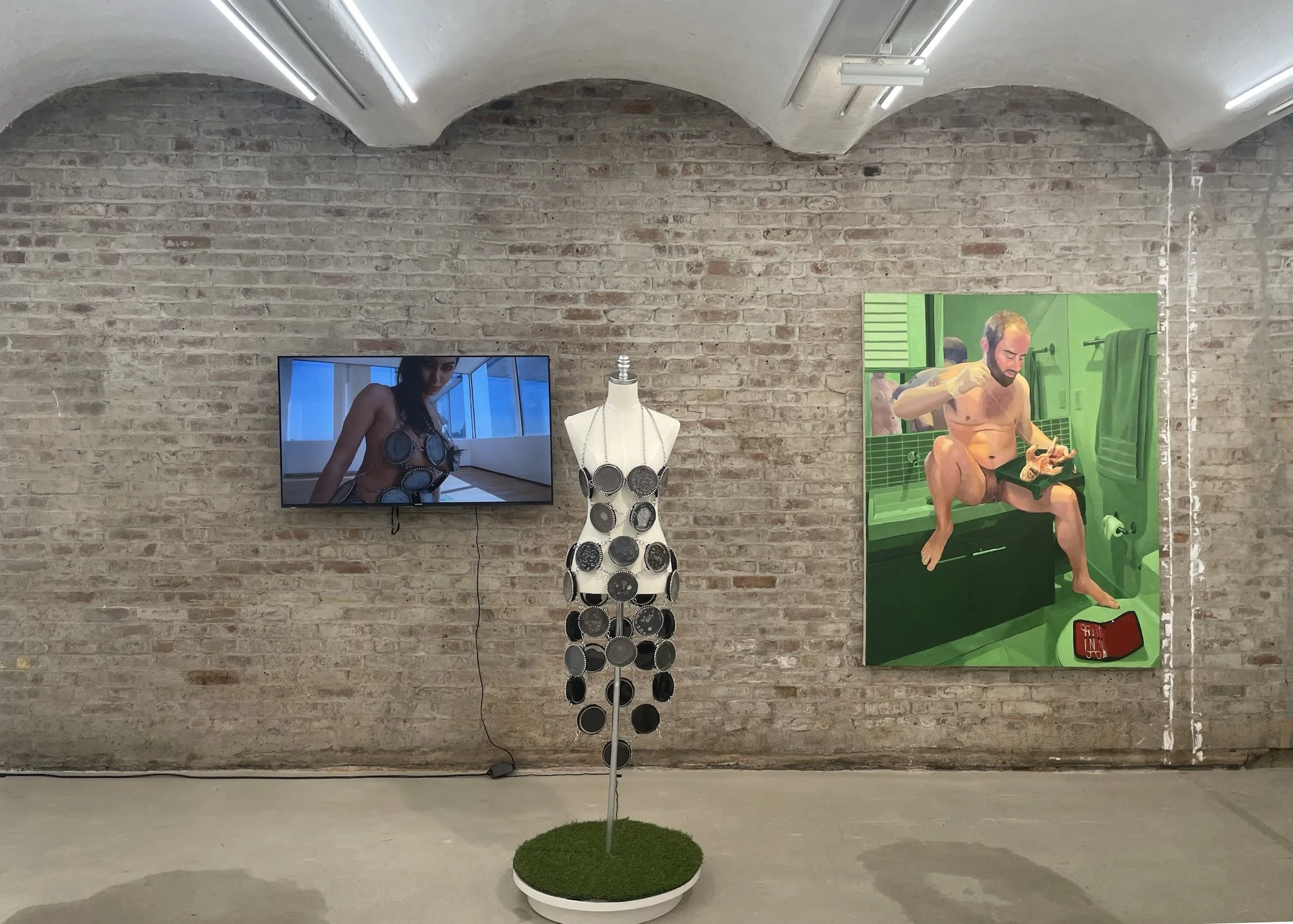 Gallery installation view with a video screen, a mannequin wearing circular metal elements, and a large green-toned painting on a brick wall.