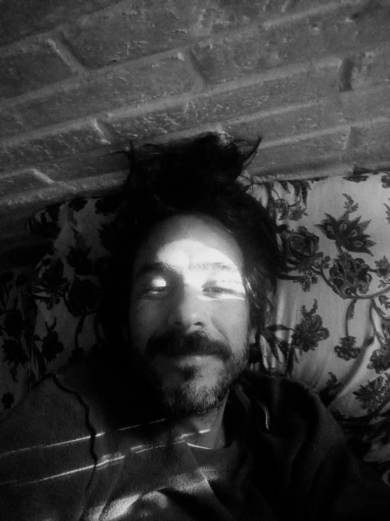 Bearded man lying down with streaks of light across his face, smiling softly in a black-and-white portrait.