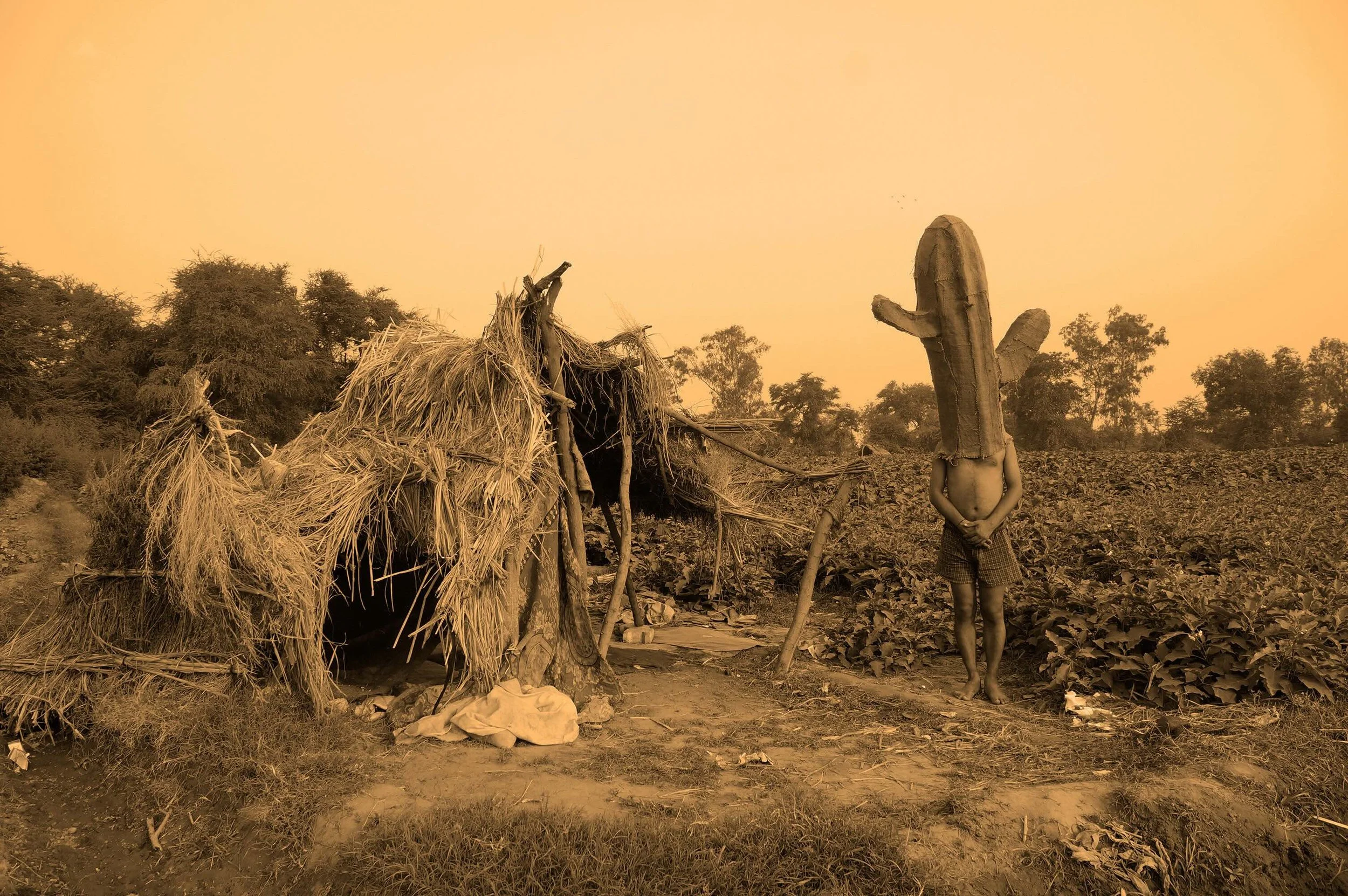 A sepia-toned photograph depicts a landscape with a dilapidated straw structure. A child stands next to the structure with a woven cactus sculpture placed over their head.