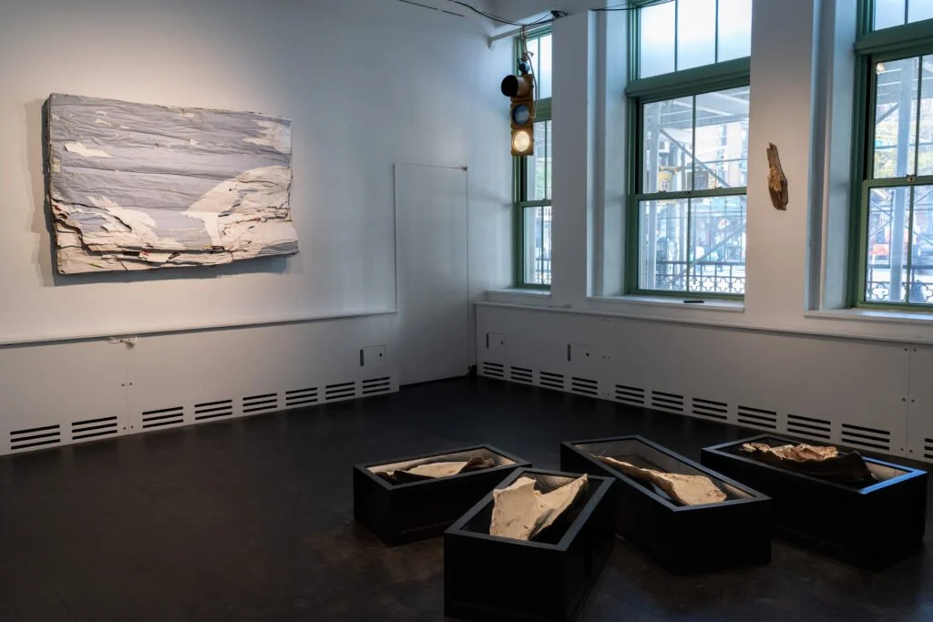 Installation view of exhibition with wall-mounted relief sculpture, hanging traffic light, and coffin-like crates on the floor.